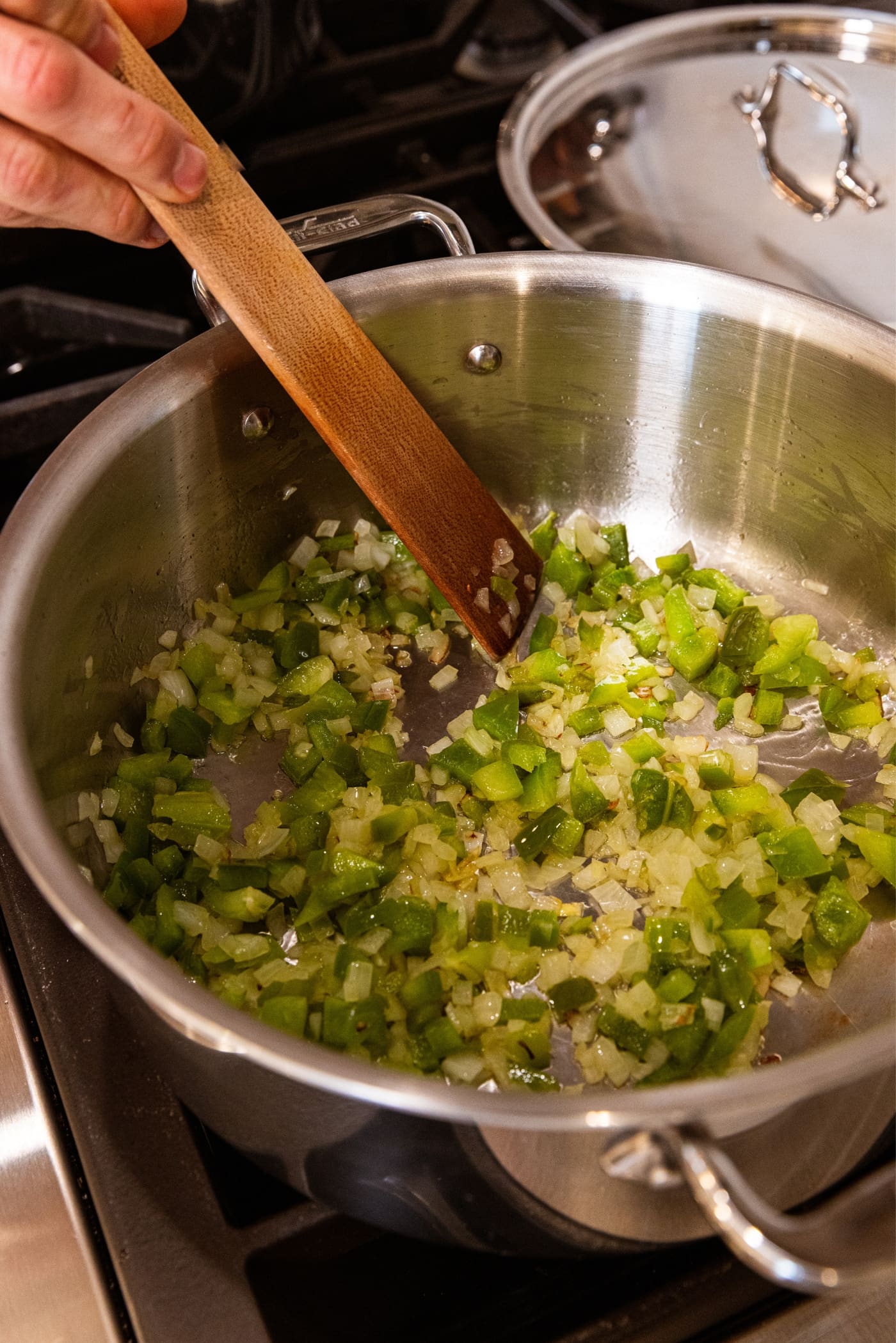 Hand holding wooden spatula mixing together browned and translucent onion and green bell pepper in metal stockpot on stovetop.