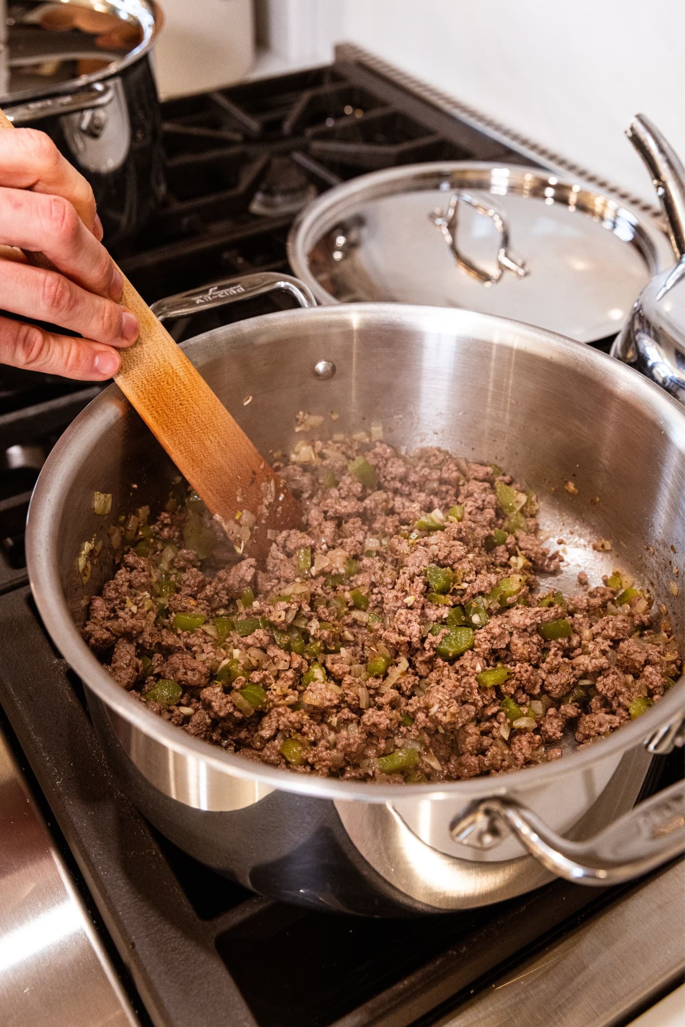 Browned ground beef in metal stockpot on stovetop with hand holding wooden spatula mixing around the cooked meat.