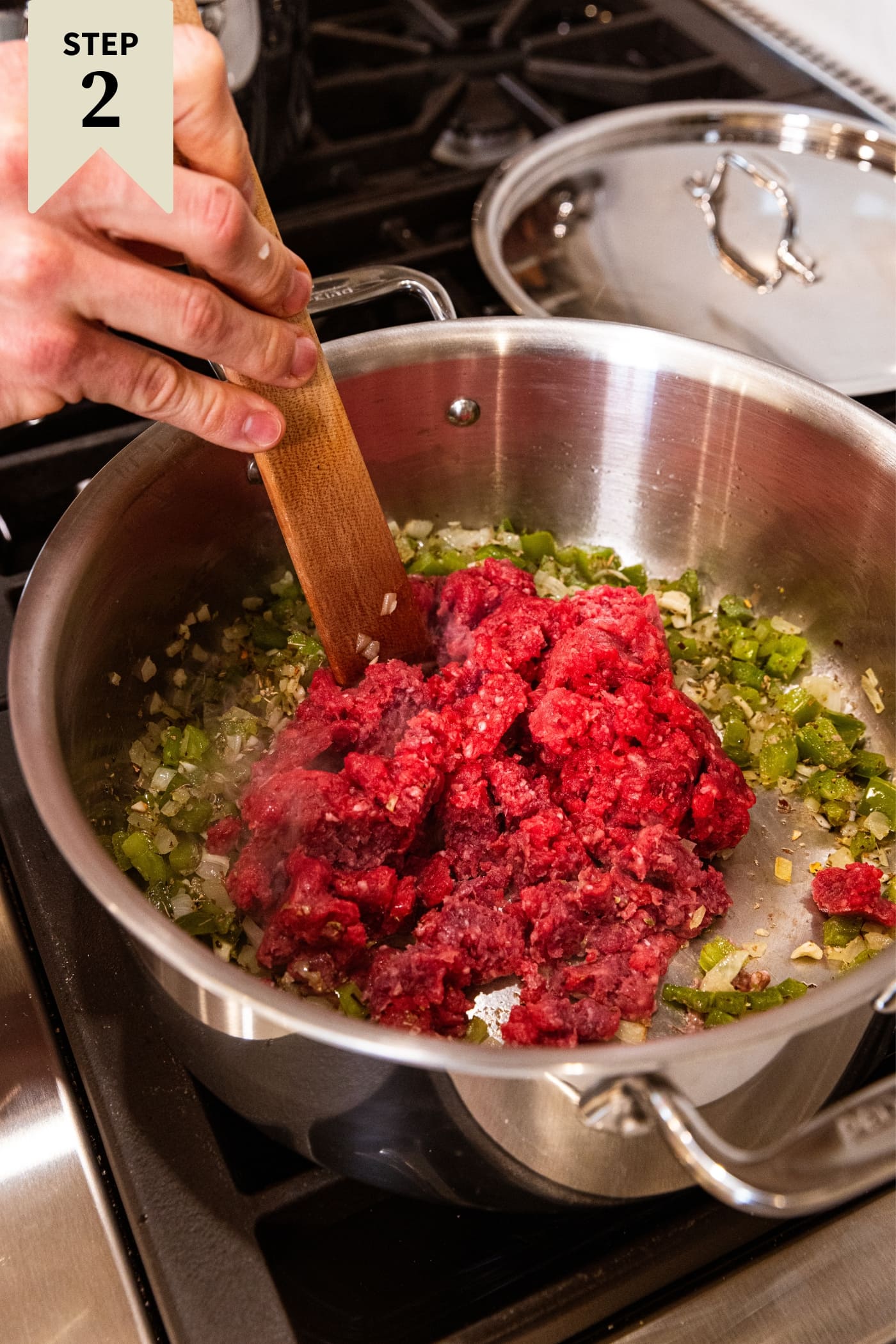 Raw ground beef sitting on top of sautéed onions and green bell peppers with hand holding wooden spatula in metal stockpot on stovetop.