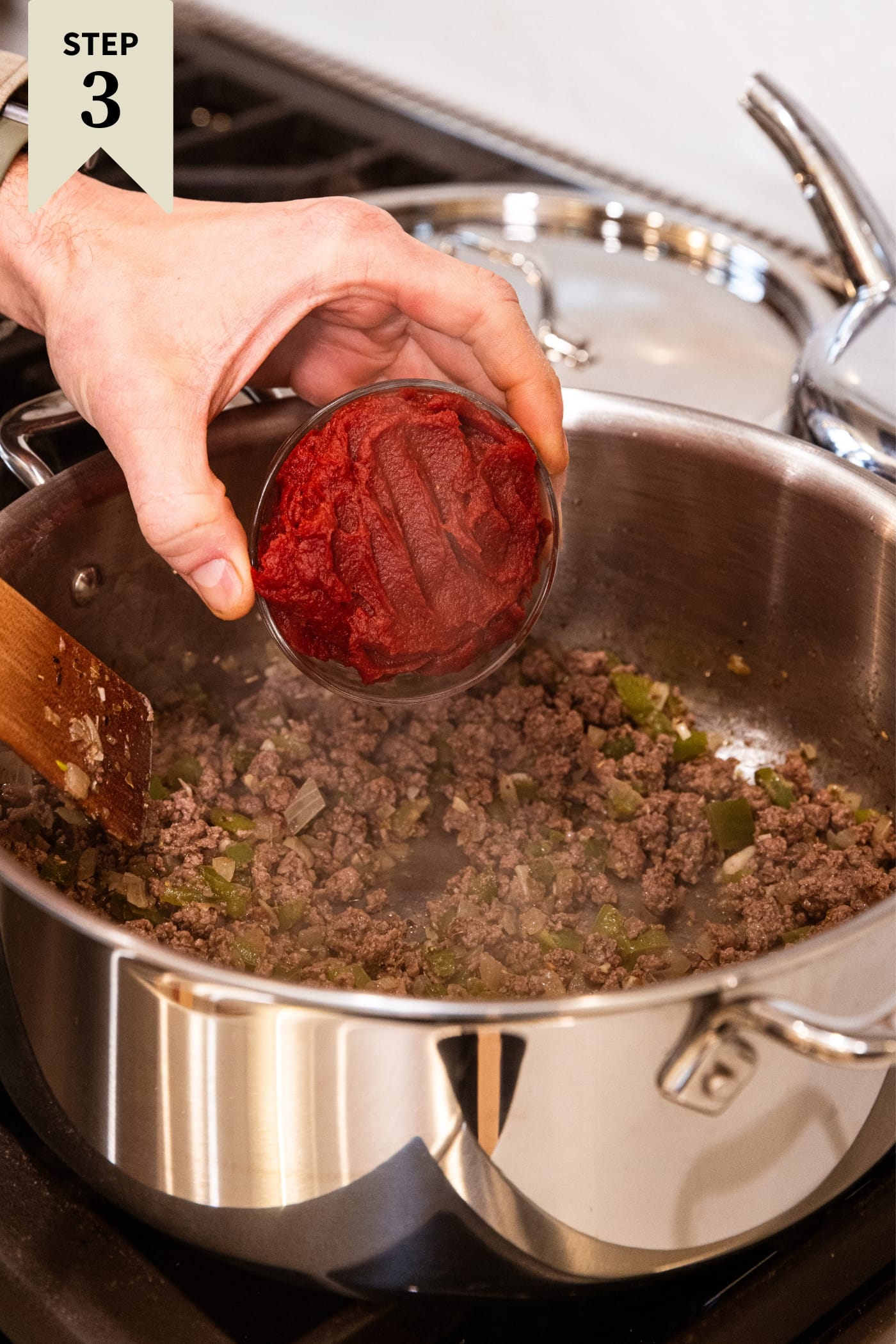 Hand holding small glass bowl filled with red tomato paste about to dump it into a stockpot filled with browned ground beef on stovetop.