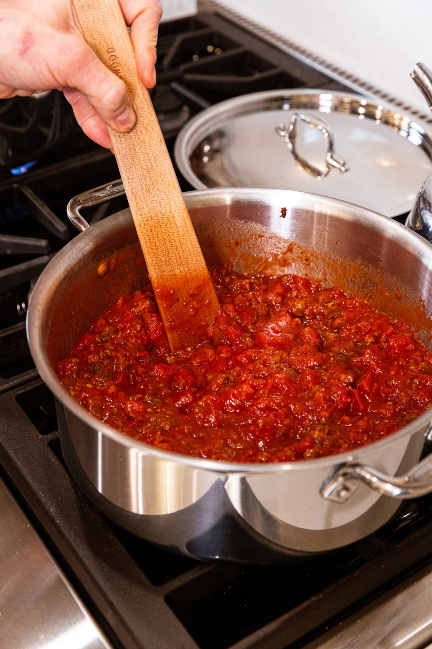 Hand holding wood spatula stirring together tomato juice and diced tomatoes with ground beef in large metal stockpot on stovetop.