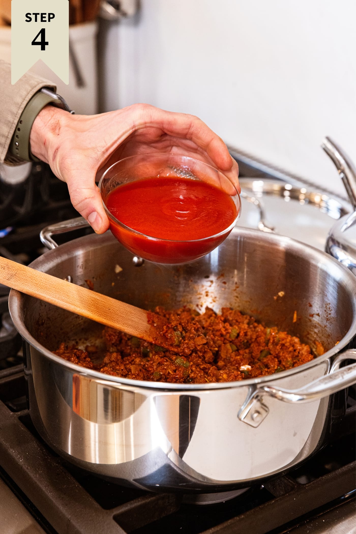 Hand holding glass bowl about to pour in tomato sauce into metal stockpot with tomato paste and ground beef mixed together.