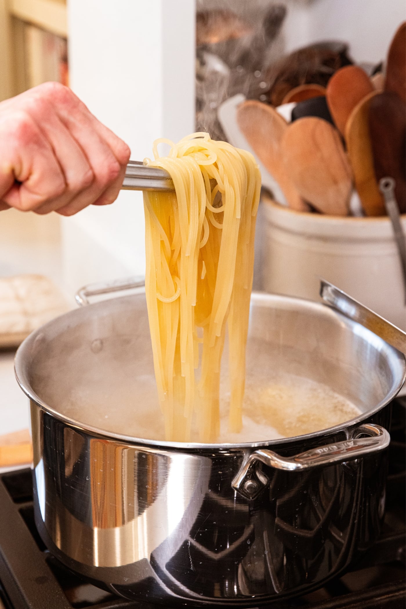 Hand holding tongs pulling up cooked spaghetti from large metal stockpot on stovetop.