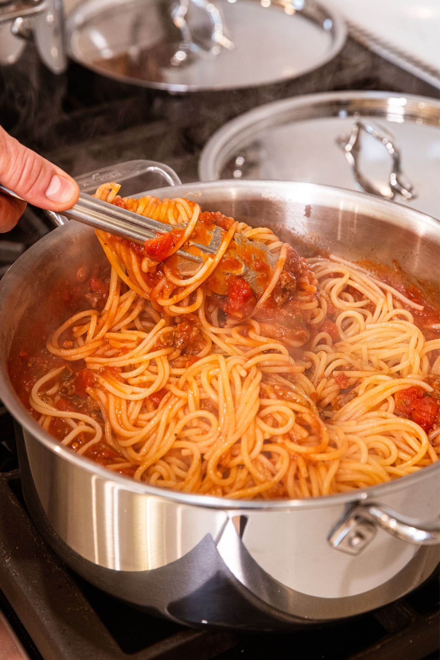Tongs holding serving of spaghetti topped with meat sauce after twisting around in metal stockpot on stovetop.