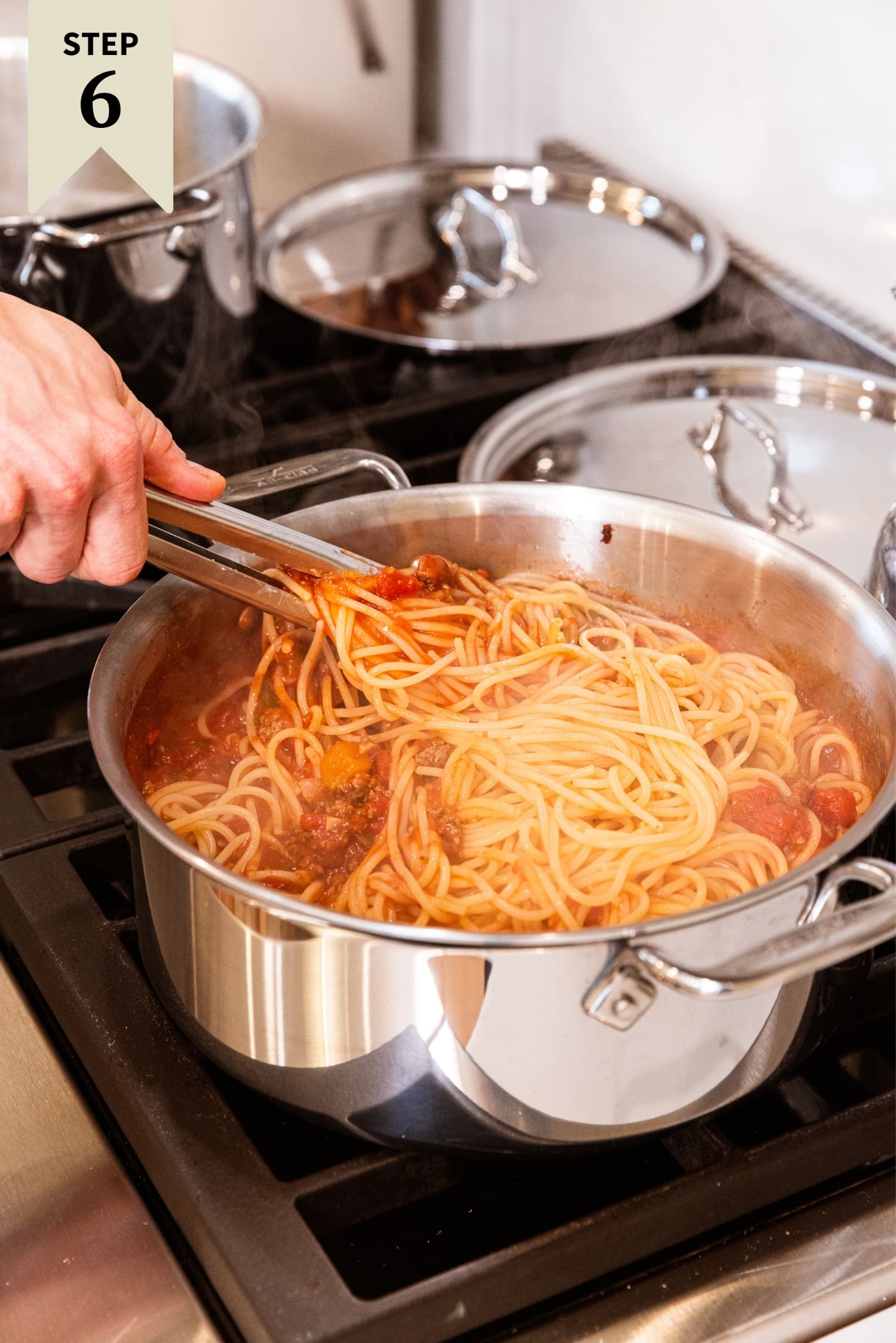 Hand holding metal tongs reaching into large metal stockpot picking up a serving of spaghetti and mixing it into the meat sauce.