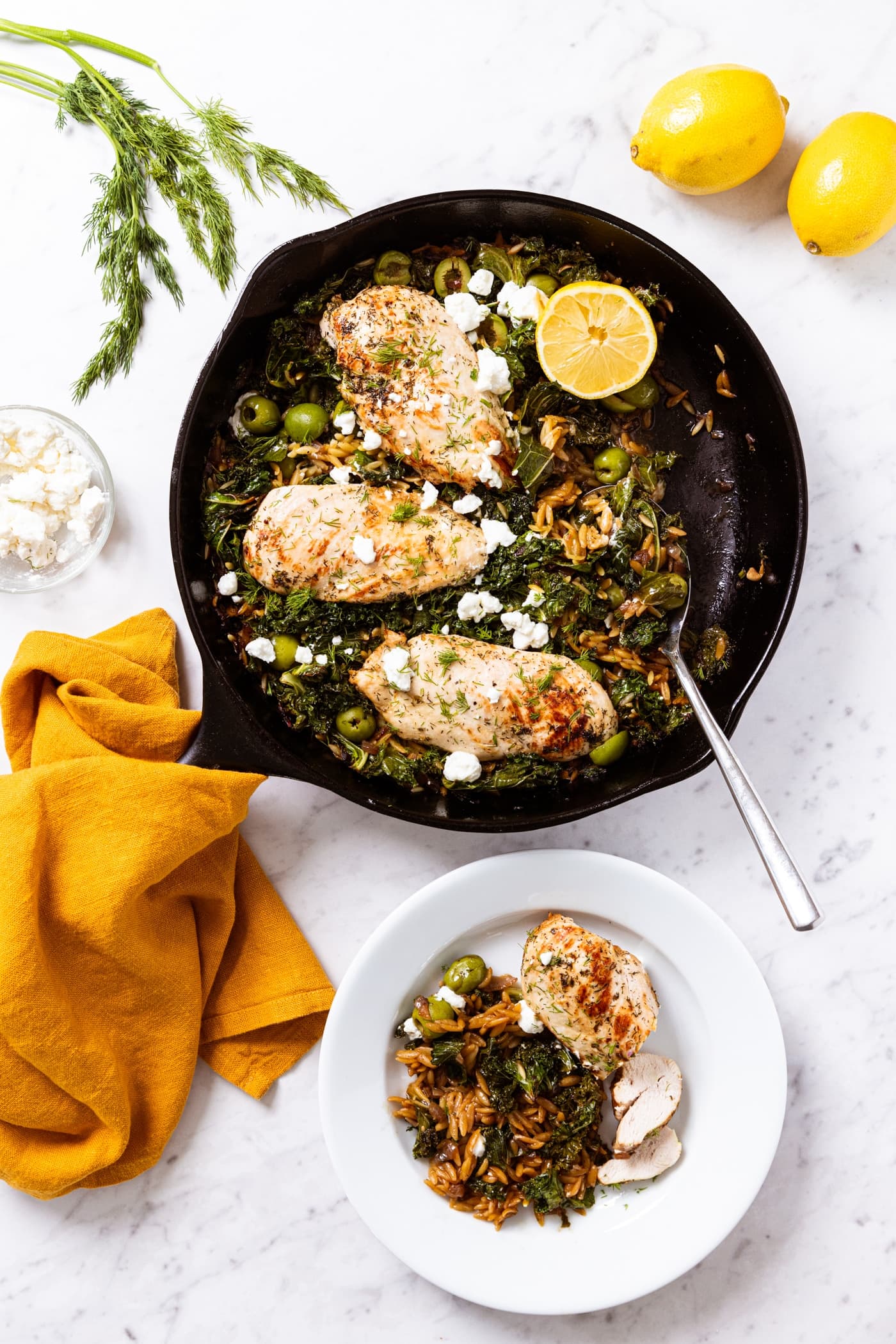 White marble surface with black cast iron skillet filled with lemon chicken with wilted kale and orzo underneath topped with crumbled feta with yellow towel holding the handle of skillet.
