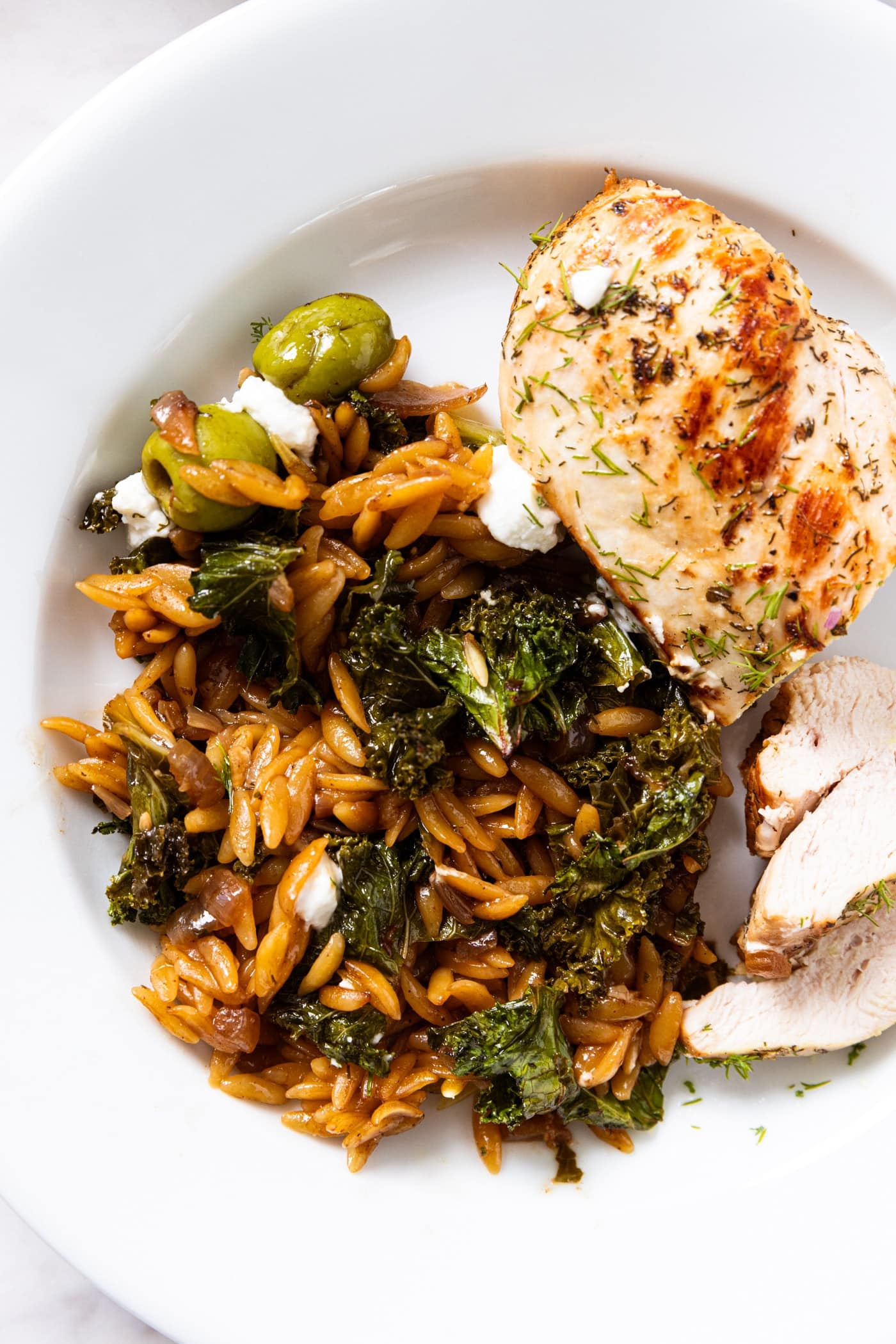 White plate filled with serving of lemon chicken with a side of cooked orzo and wilted kale topped with crumbled feta cheese.