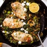 Black cast iron skillet sitting on marble surface filled with cooked chicken breasts and orzo with half of lemon nestled amongst wilted kale with spoon resting on side of skillet.