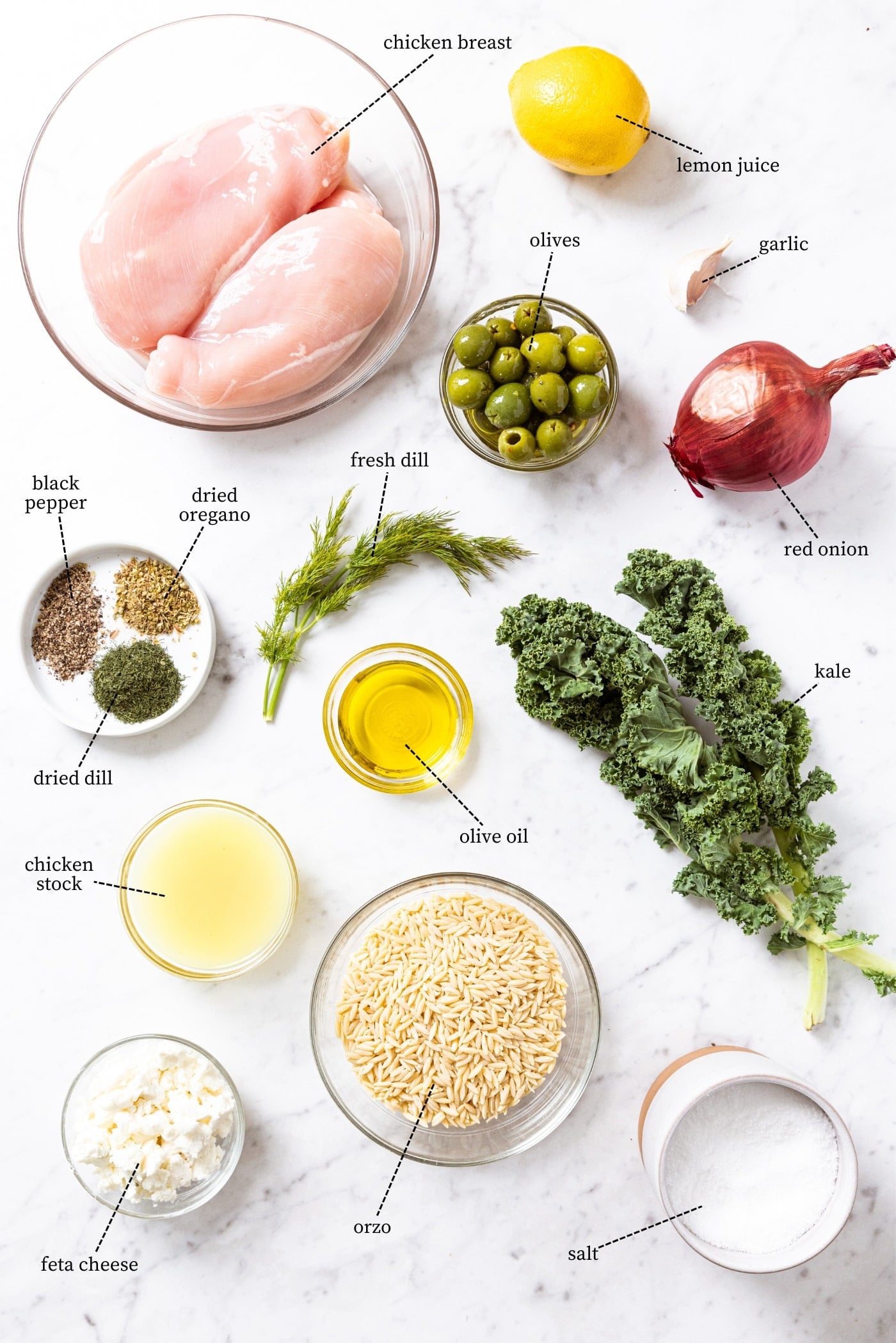 White marble surface filled with all ingredients needed to make lemon chicken including kale, red onion, olives, chicken breasts, orzo, spices, and more.
