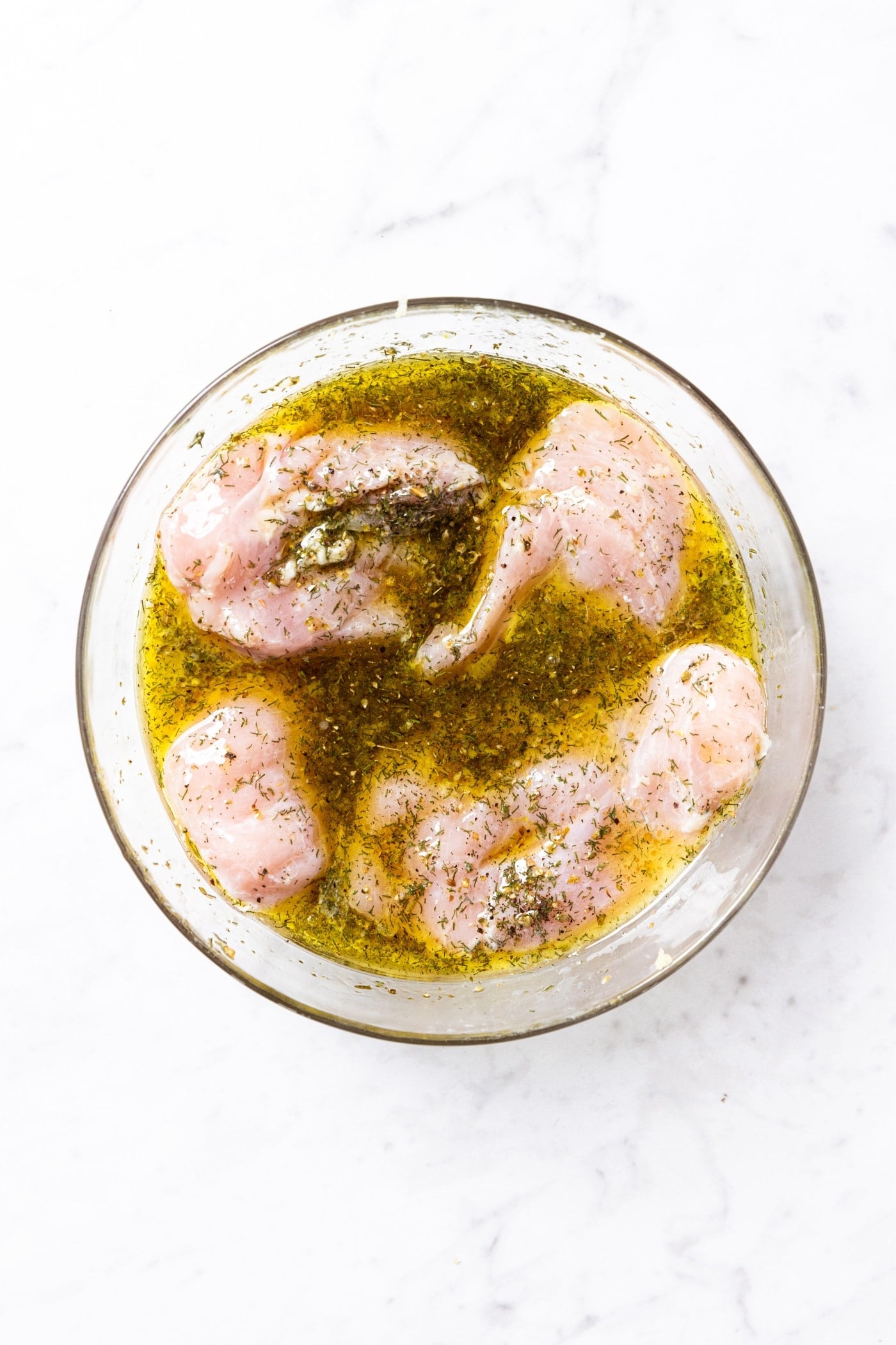 Glass bowl sitting on white surface filled with marinated raw chicken breasts covered with spices and olive oil mixture.