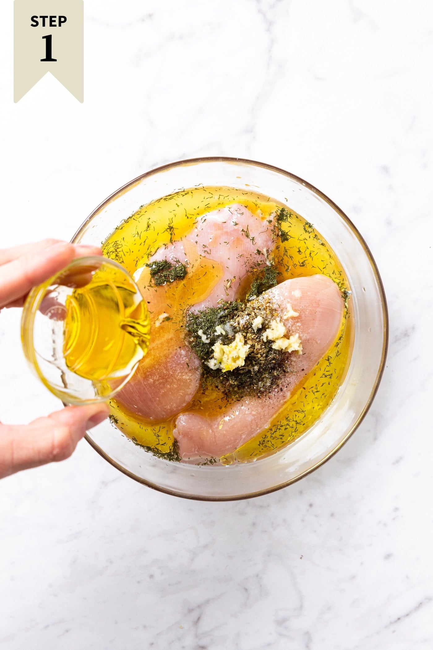Glass bowl with raw chicken breasts inside with spices, garlic, and lemon juice with hand holding smaller glass bowl pouring olive oil on top.