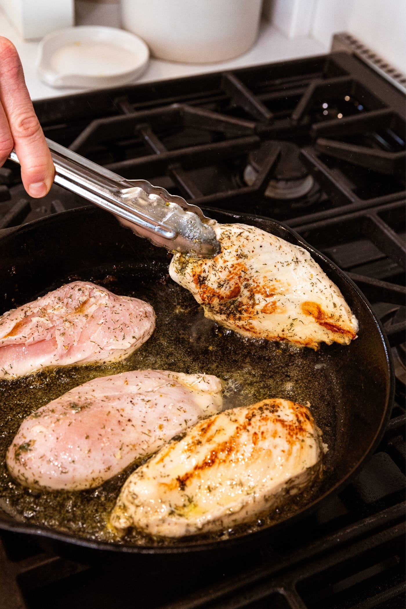 Hand holding tongs lifting half-cooked chicken breast out of black cast iron skillet on stovetop with other chicken breasts in pan as well in varying states of cooking.