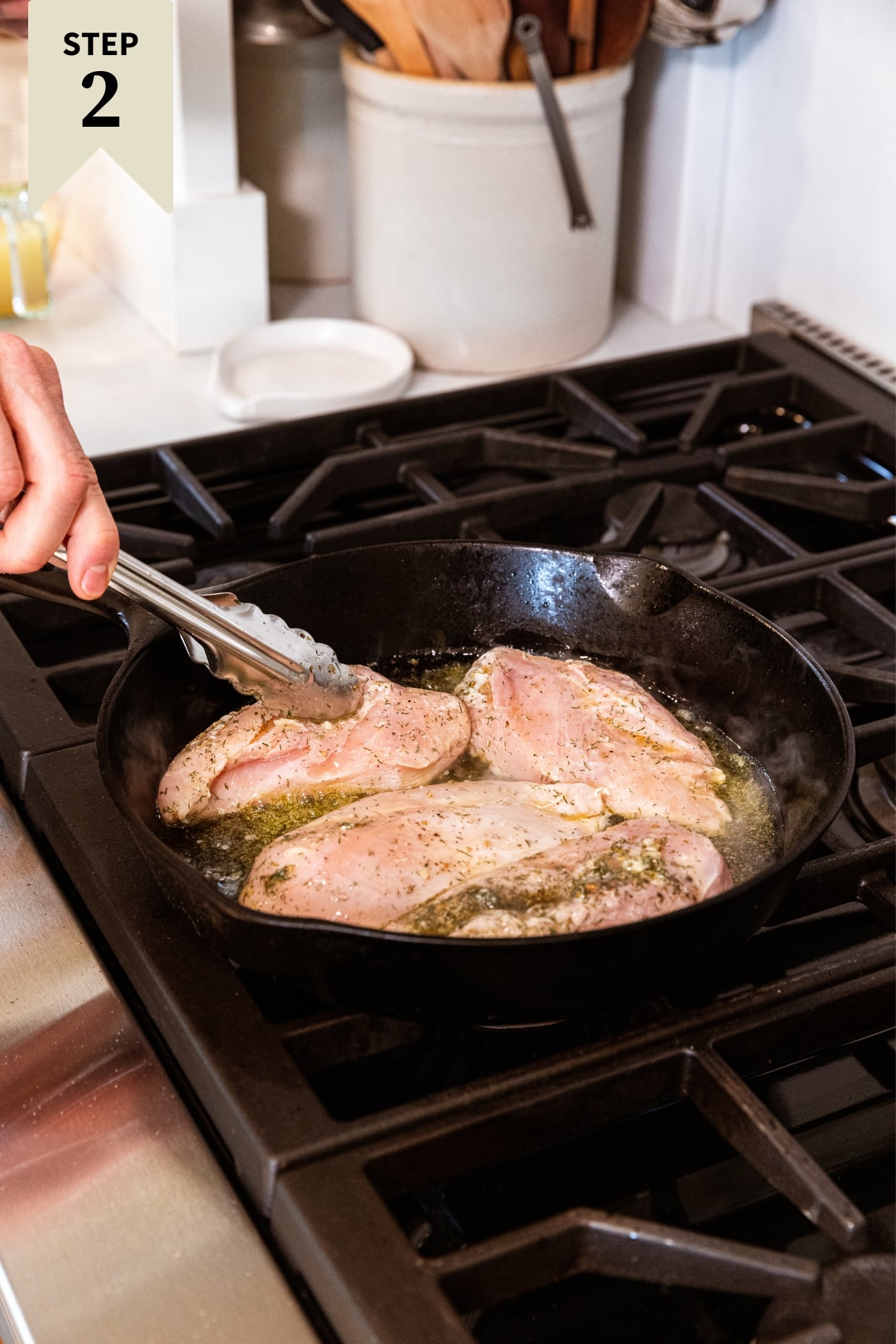 Hand holding tongs placing raw chicken breasts in black cast iron skillet filled with oil all on stovetop.