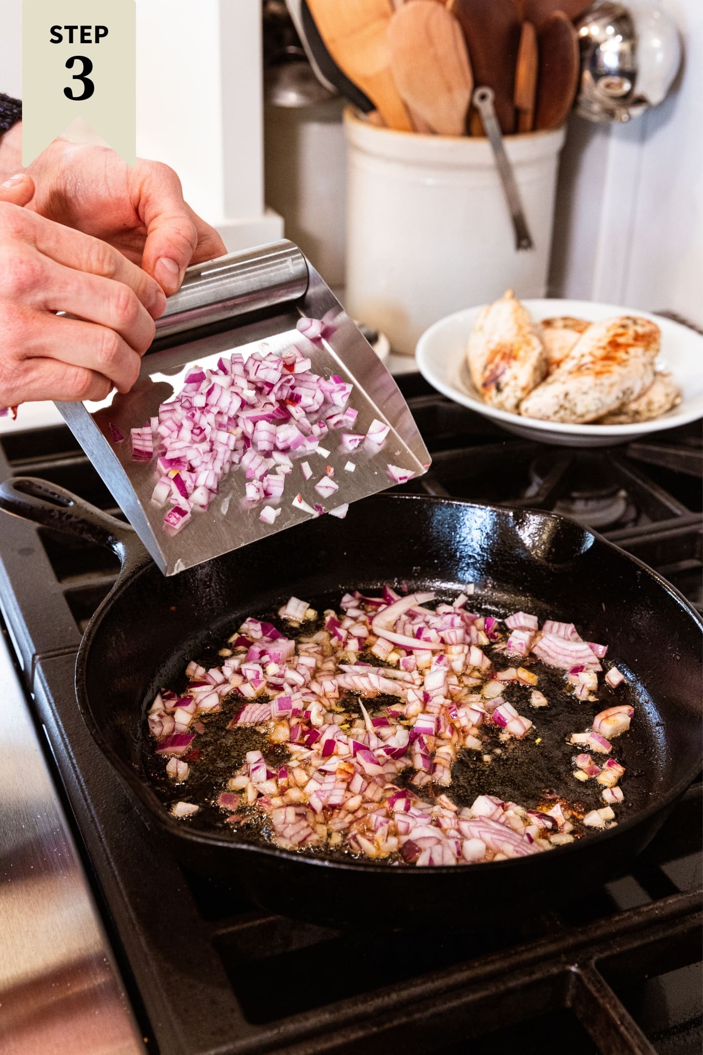 Hand using metal scooper to pour chopped red onion into black cast iron skillet sitting on stovetop with oil in pan with cooked chicken breasts sitting on a plate in the background.