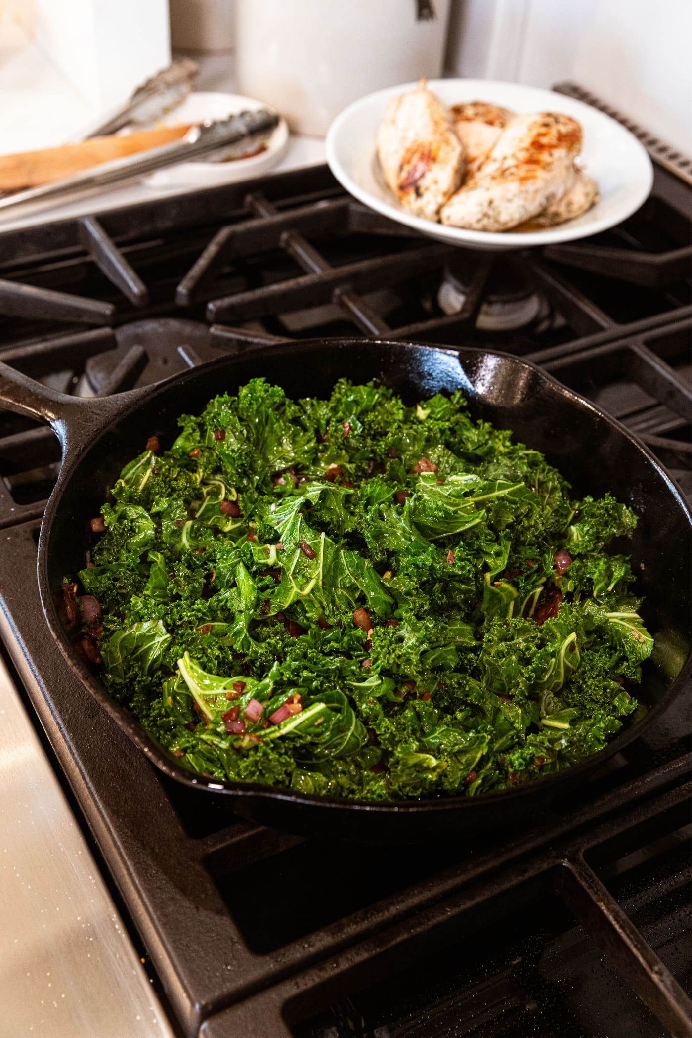 Black cast iron skillet sitting on stovetop filled with wilted green-colored kale mixed with browned red onion with white plate filled with cooked chicken breasts in background.