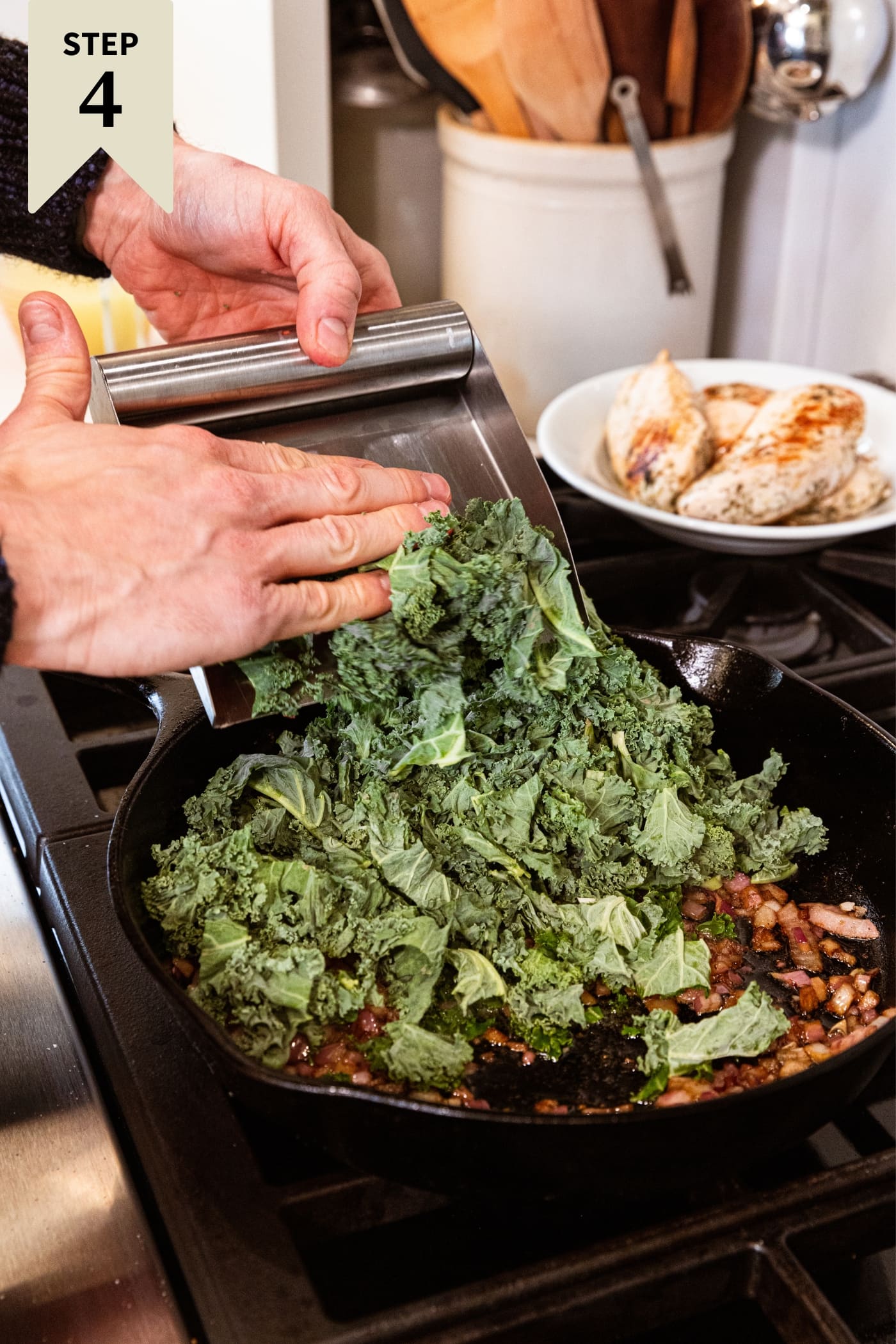Hand scooping chopped raw kale into a black cast iron skillet on stovetop from metal scooper with skillet already filled with browned red onion.
