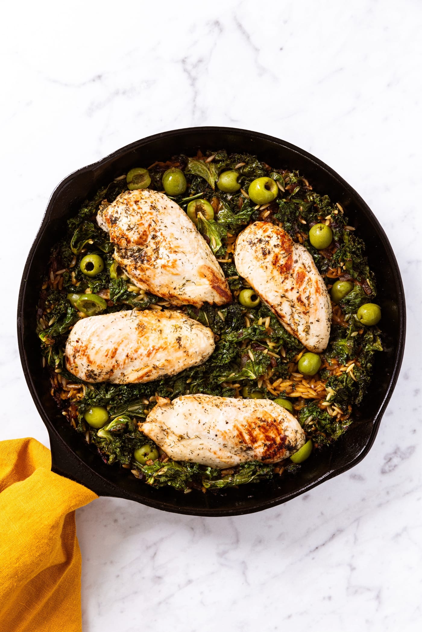 White marble surface with black cast iron skillet filled with cooked chicken breasts sitting on a bed of wilted kale, olives, orzo, and more.