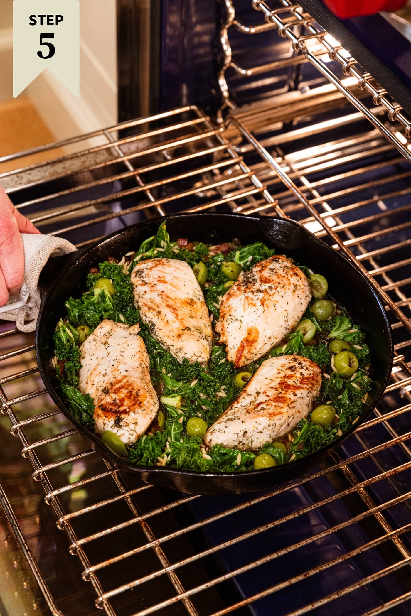 Hand holding black cast iron skillet filled with wilted kale with browned chicken breasts on top about to go into the oven to finish cooking.