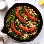 White marble surface with black skillet filled with browned sausages on top of white beans mixed with wilted kale.