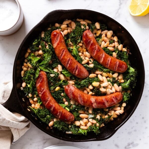 White marble surface with black skillet filled with browned sausages on top of white beans mixed with wilted kale.