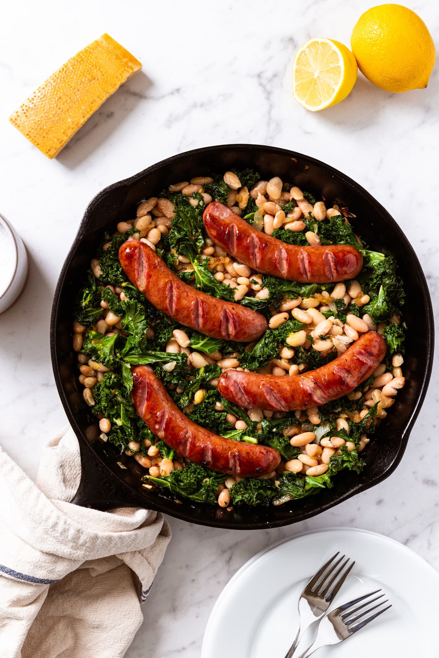 White marble surface with black skillet filled with browned sausages on top of wilted kale with white beans underneath with lemons and Parmesan cheese also on marble.