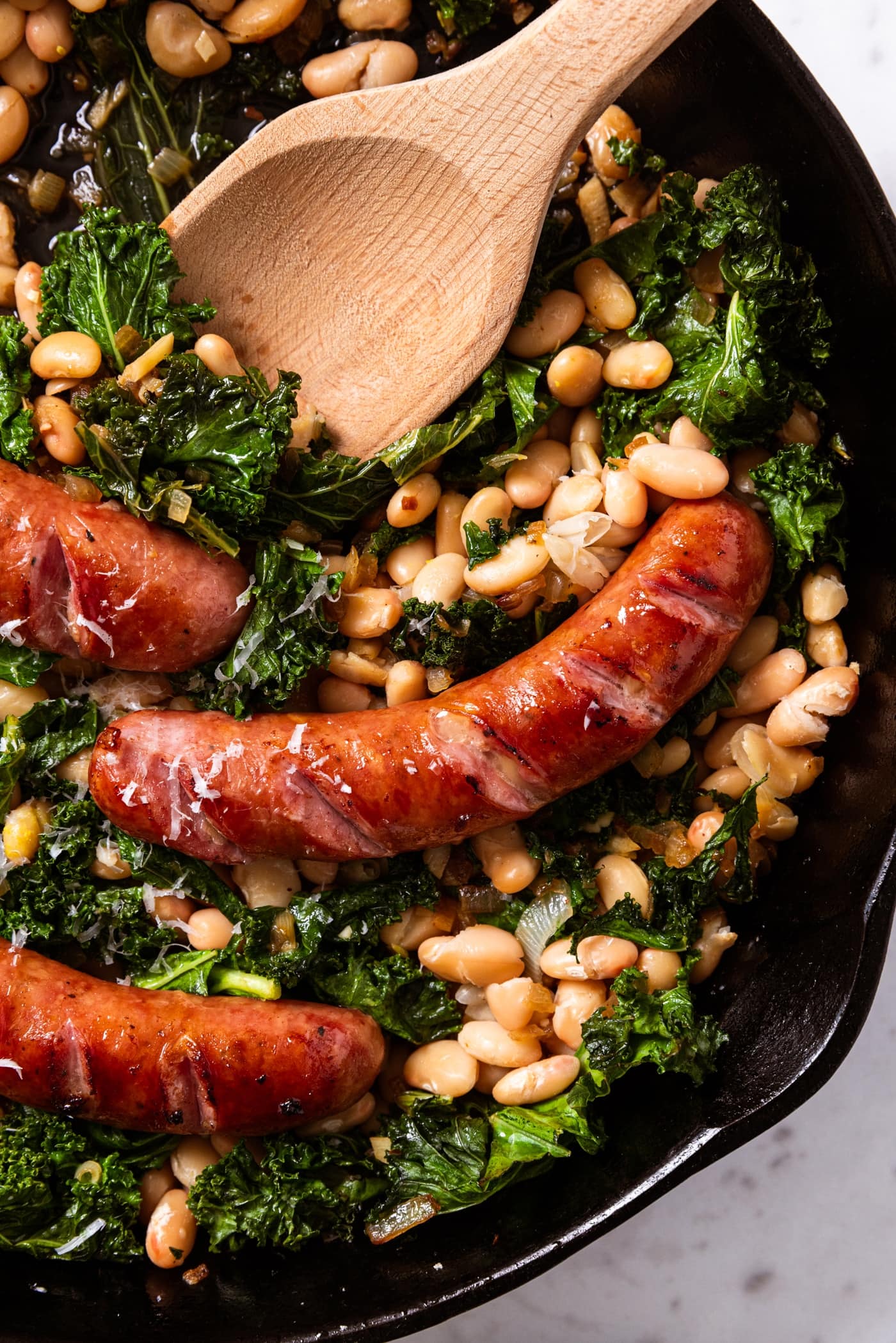 Black skillet sitting on marble surface filled with browned sausage links on top of wilted kale and white beans with wooden spoon resting on the side.