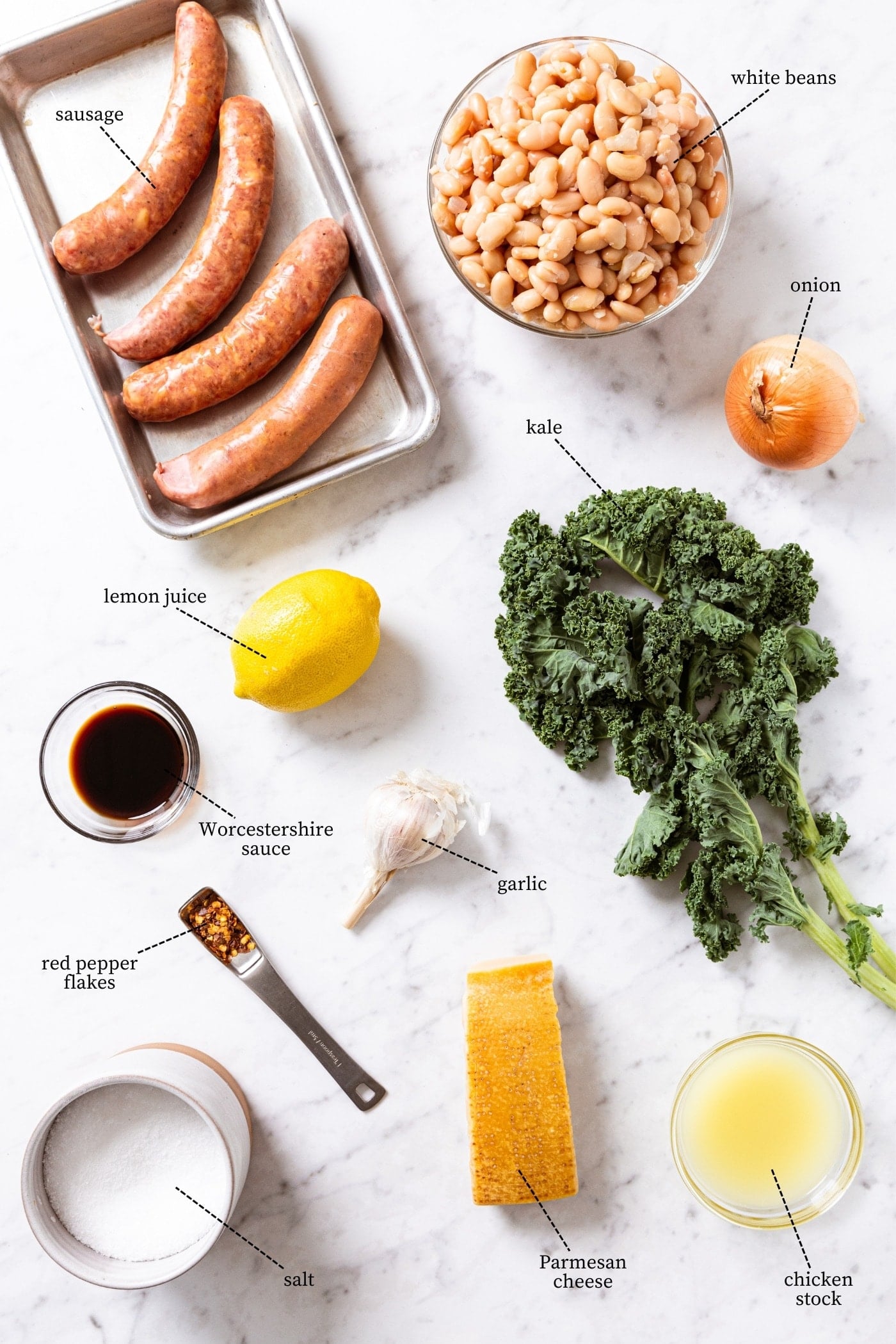 White marble surface filled with all ingredients needed to make skillet sausage with sausages, white beans, kale, onion, lemon, cheese, and more.