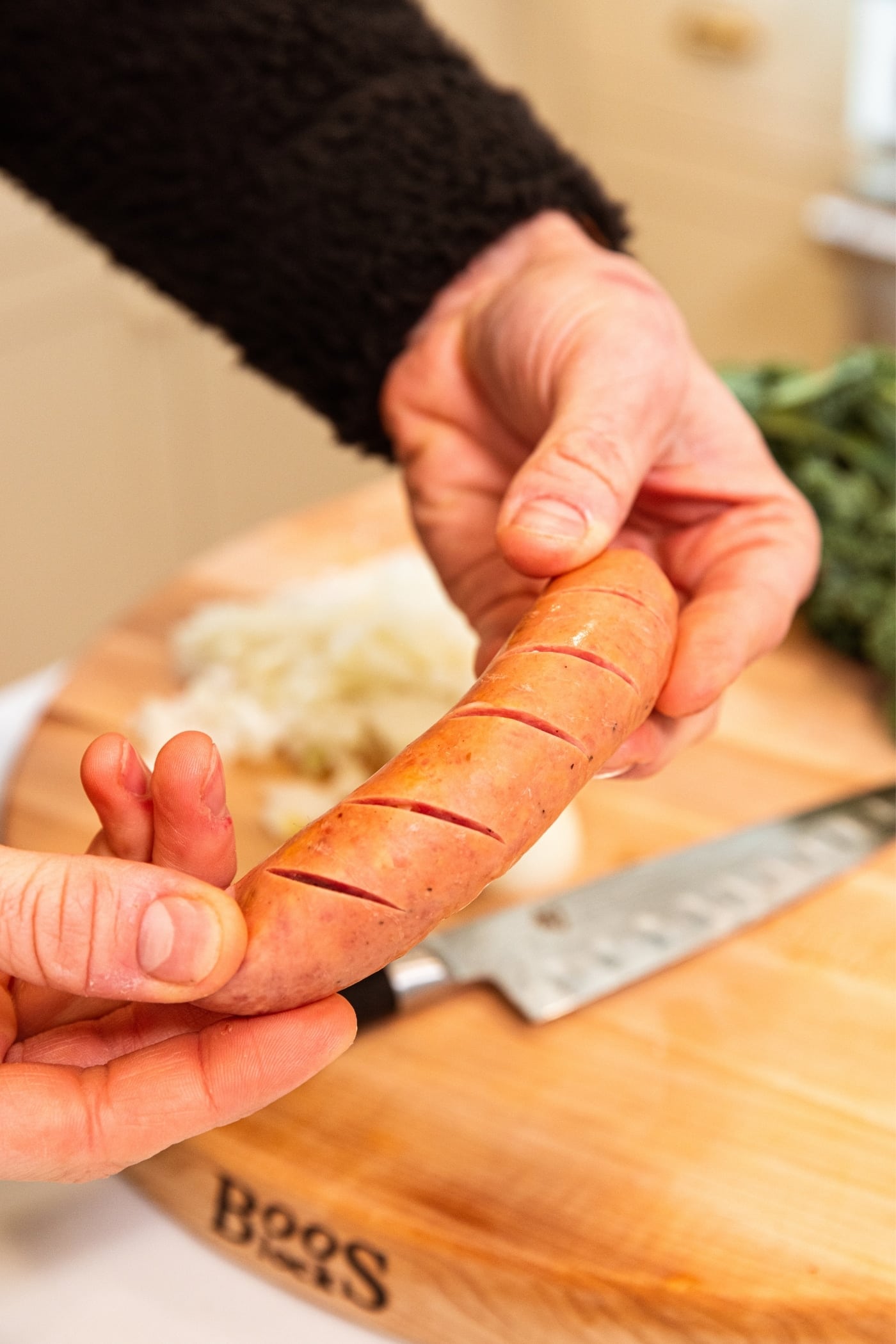 Two hands holding uncooked sausage after making small marks in sausage with knife as relief cuts with wood cutting board in background.