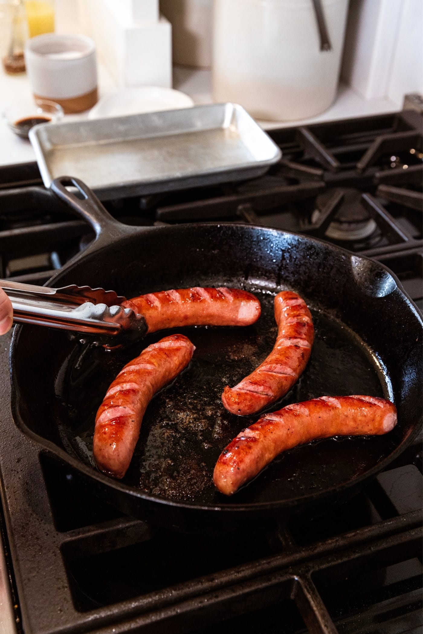 Hand holding tongs after flipping sausage links with brown marks on the top in black skillet on stovetop.