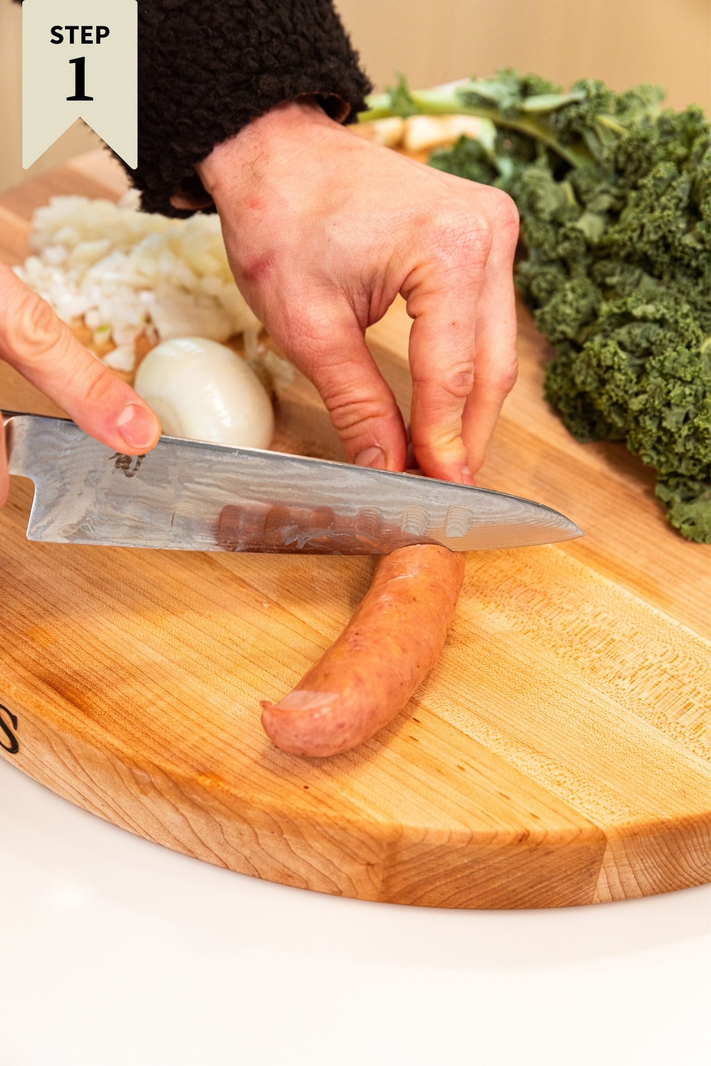 Hand holding knife creating cut marks on the top of the sausage on wood cutting board with kale and onion in background.