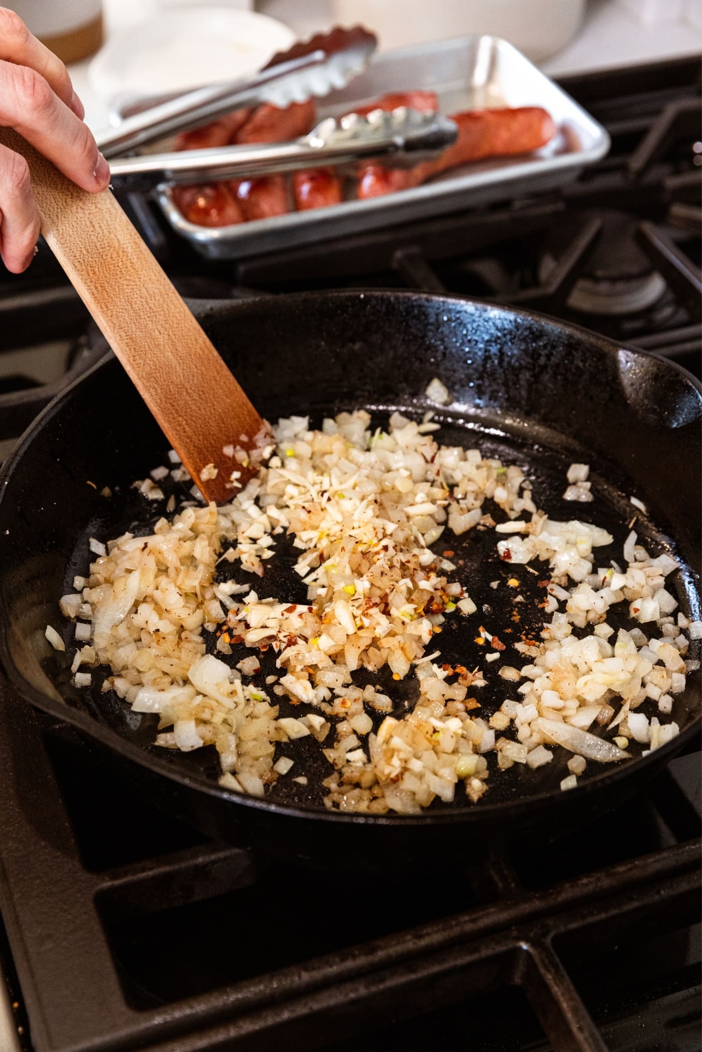 Wooden spatula scraping up browned onion and red pepper flakes in black skillet on stovetop with browned sausages in background on metal tray.