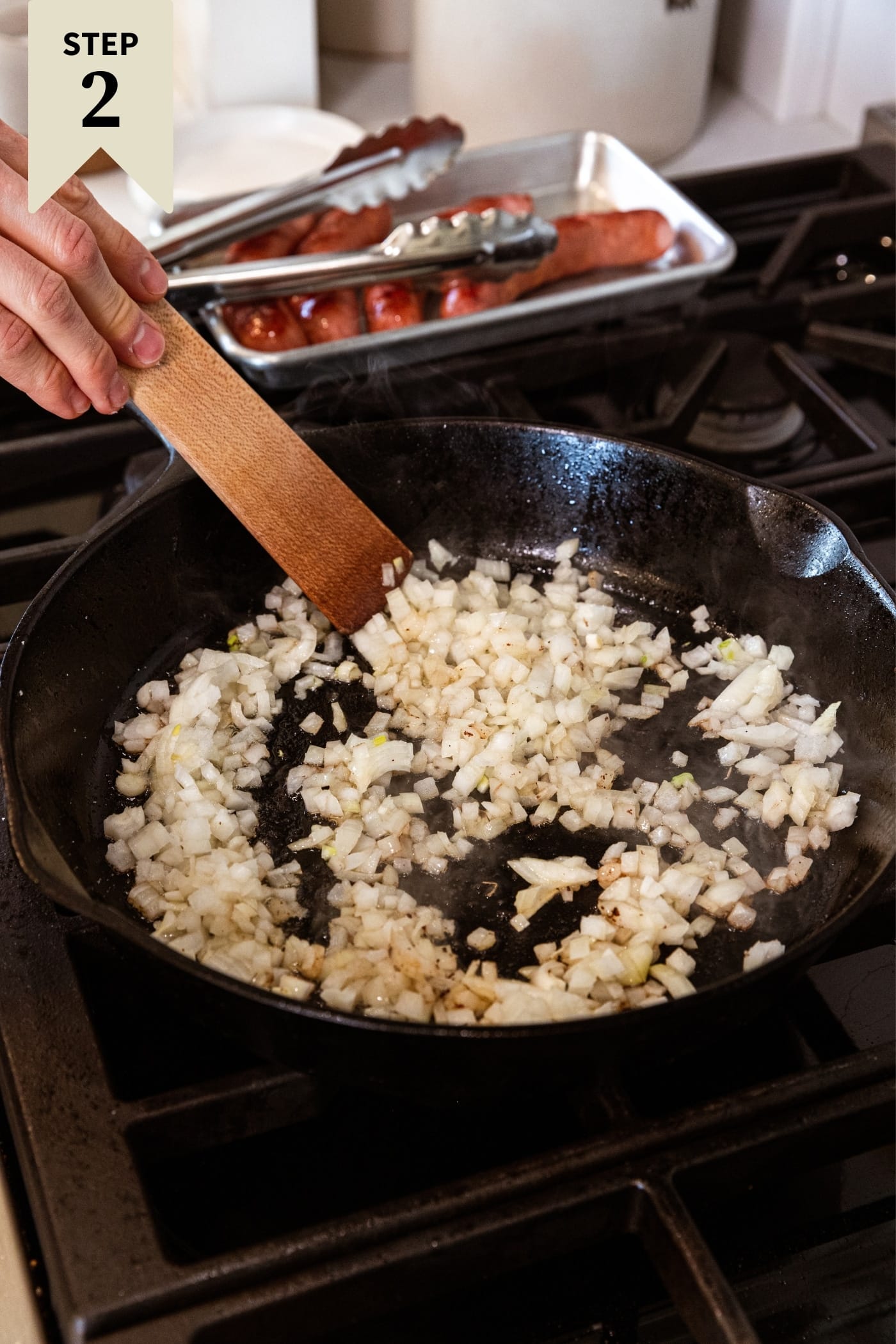 Hand holding wooden spatula scraping diced onion in black skillet on stovetop right after adding to the skillet.