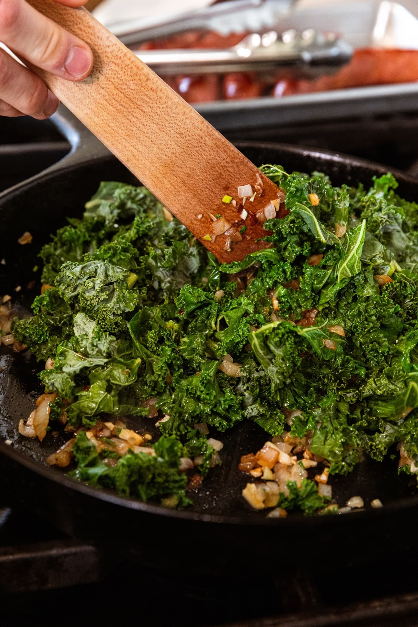 Wilted kale sitting in black skillet after sitting on heat for awhile with wooden spatula mixing everything together.