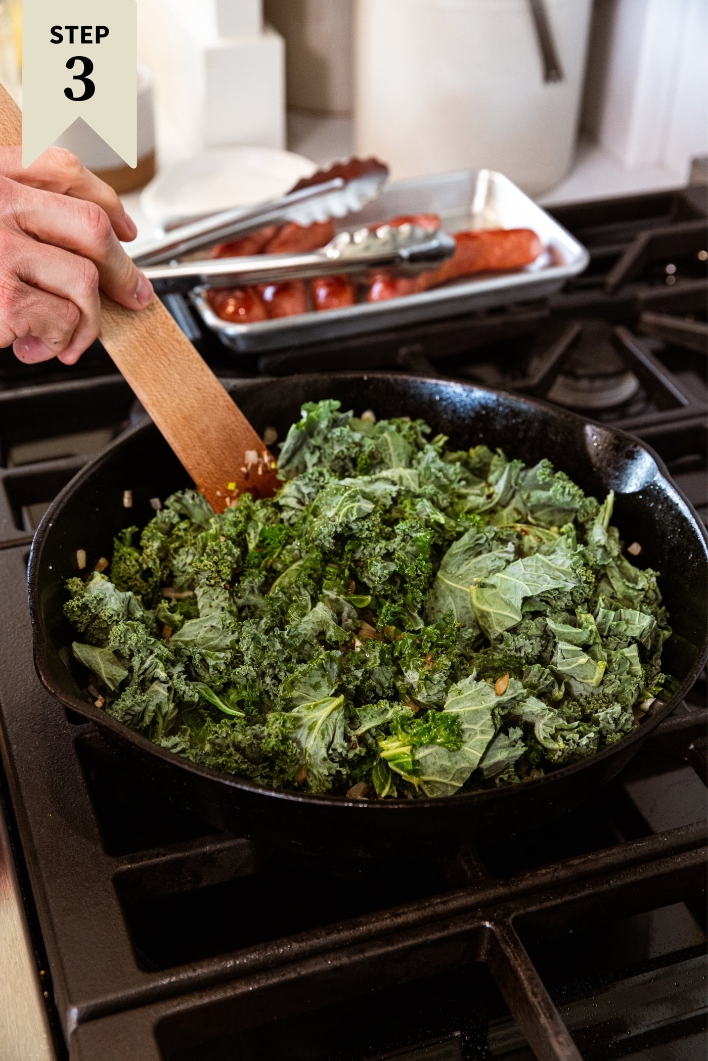 Black skillet sitting on stovetop with roughly chopped kale before being wilted over heat with browned sausage in background.