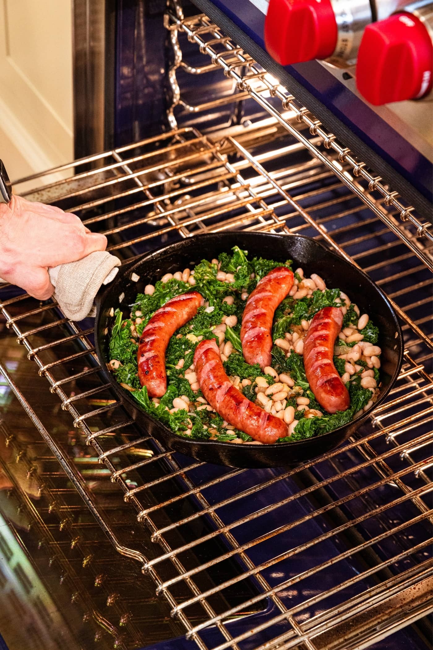 Hand placing black skillet with wilted kale and sausage on rack in oven before closing the door to finish baking it.