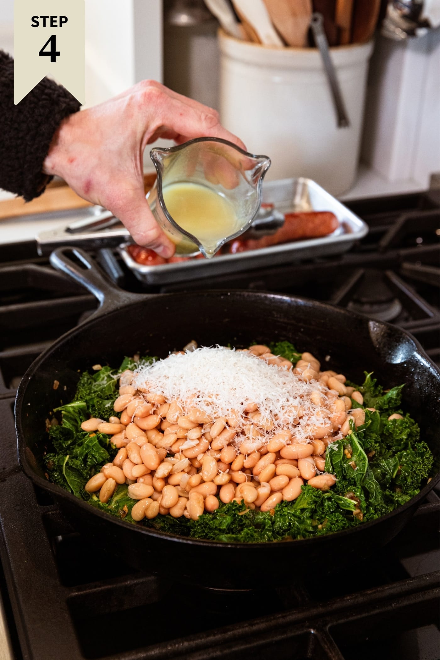 Hand holding glass measuring cup about to pour chicken stock on top of white beans and shredded Parmesan cheese sitting in black skillet.