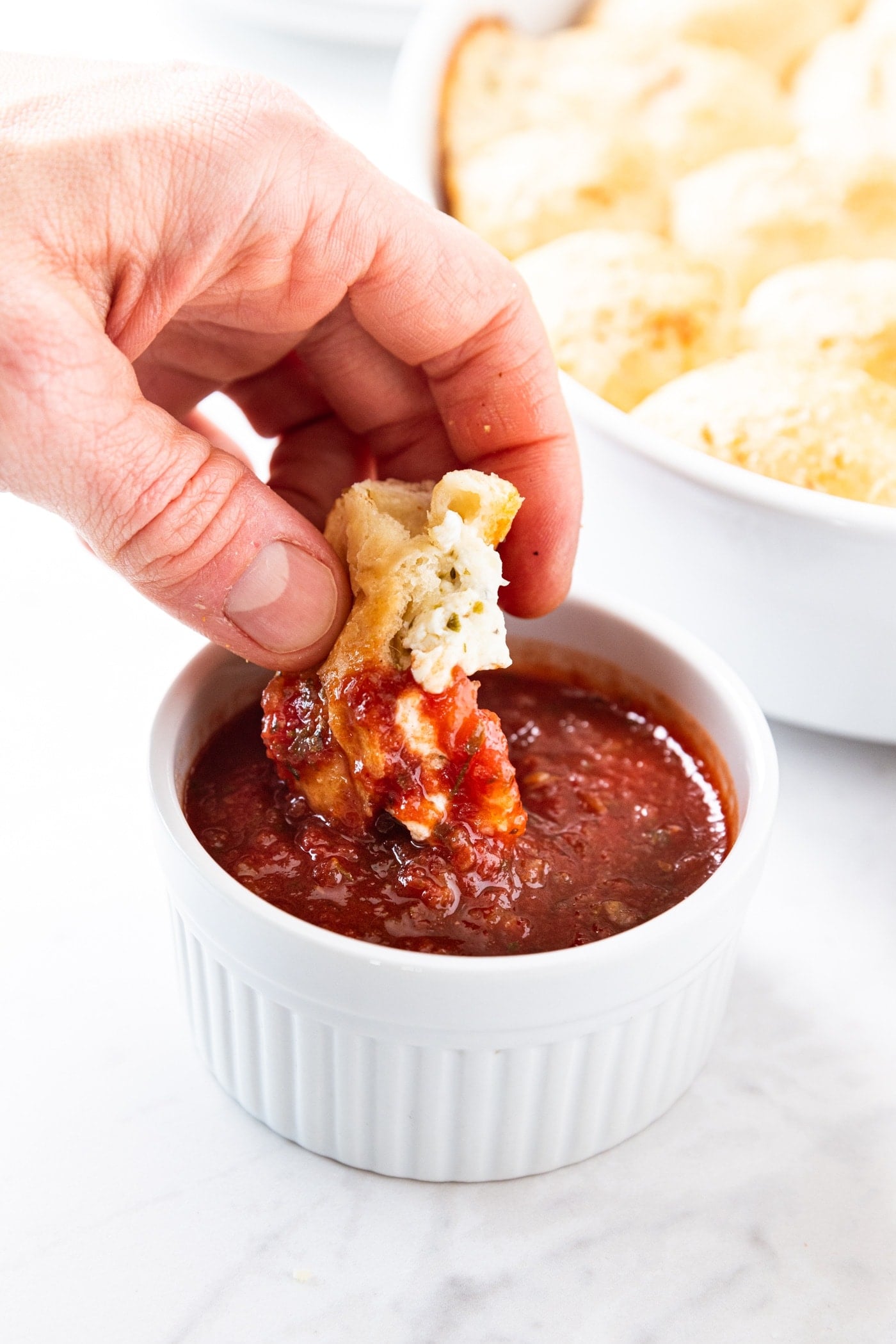 Hand holding cheese pizza bun dipping into a small white bowl filled with marinara sauce.