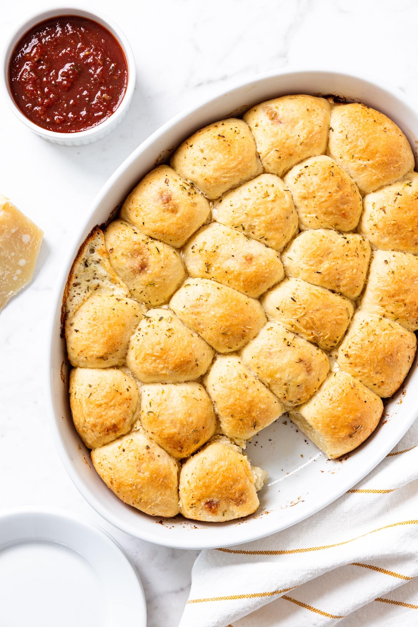 Marble surface with white oval baking dish filled with baked pizza bites with small white bowl filled with marinara dipping sauce to the side.