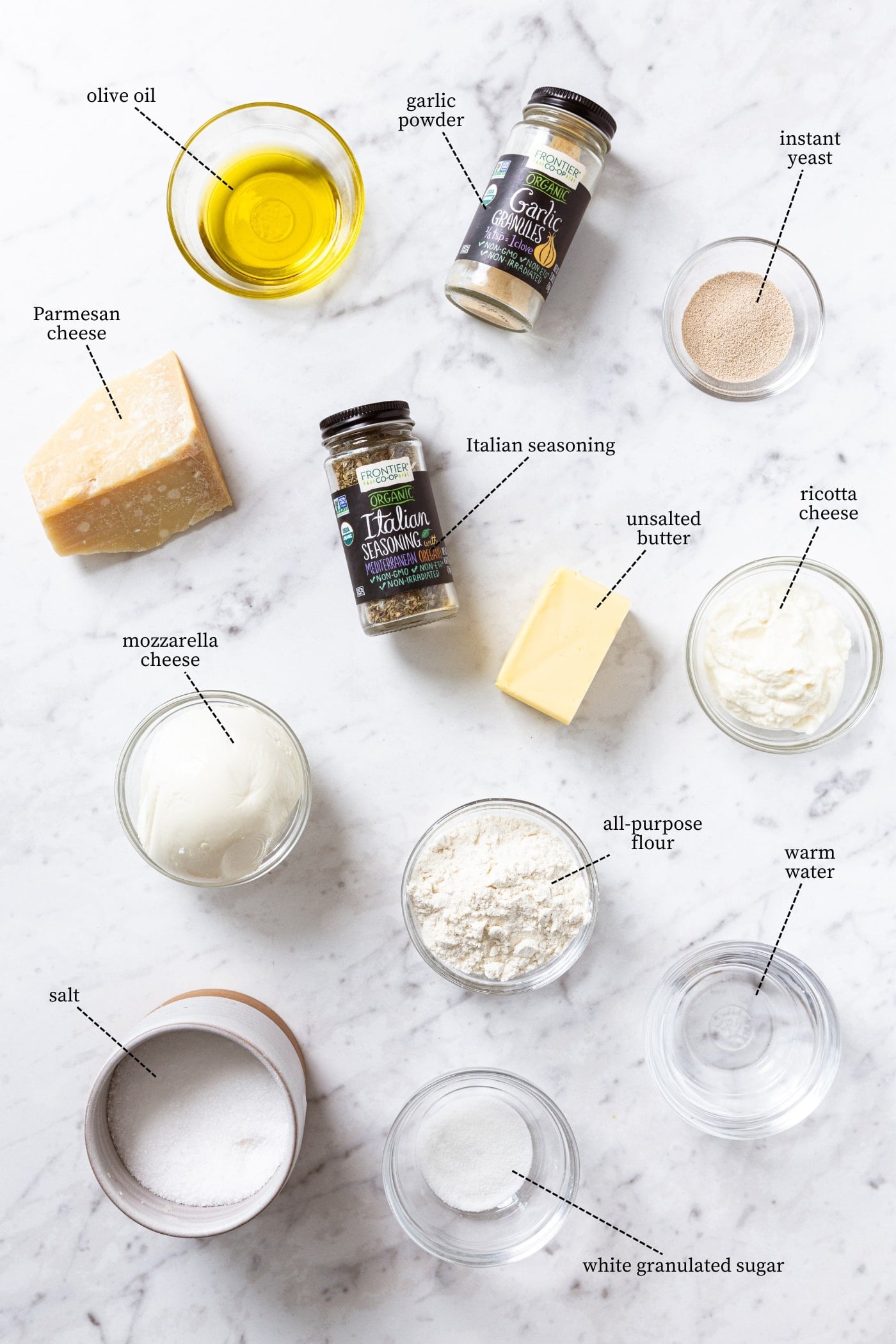 White marble surface filled with all ingredients needed to make pizza bites including Parmesan cheese, ricotta cheese, flour, salt, butter, spices, and more.