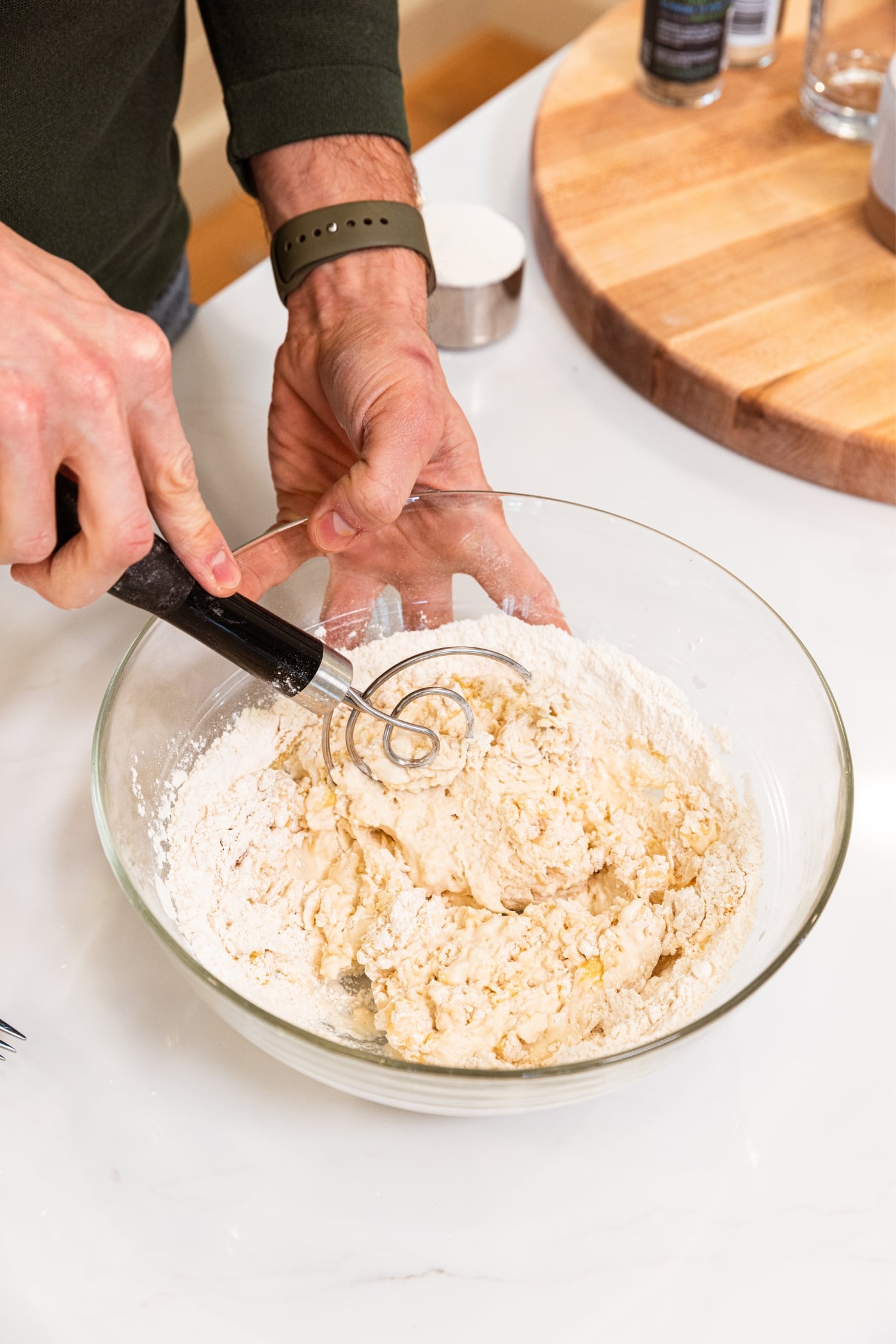 Glass bowl with flour mixture with hand mixing together everything with wire whisk on white countertop.
