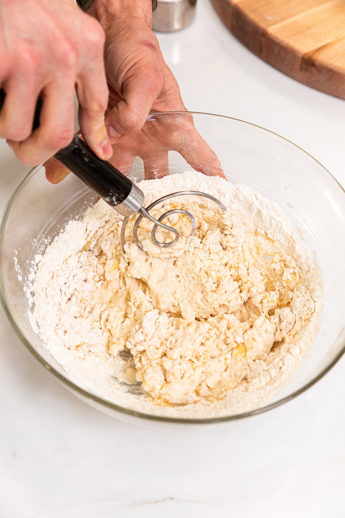 Wire dough whisk being held whisking together flour with wet ingredients in large glass bowl.