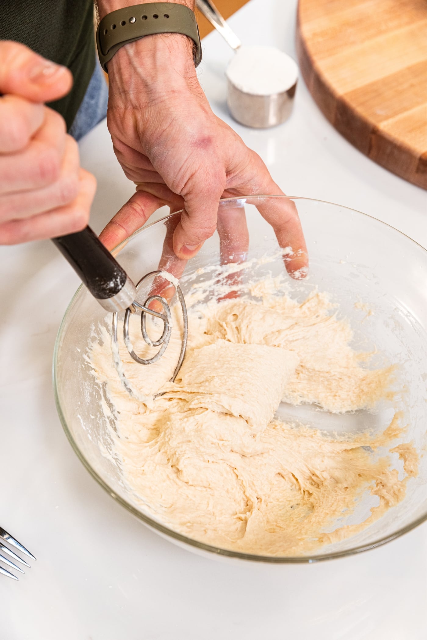 Hand pulling dough to the side of a glass bowl with wire dough whisk on white countertop showing texture of the dough.