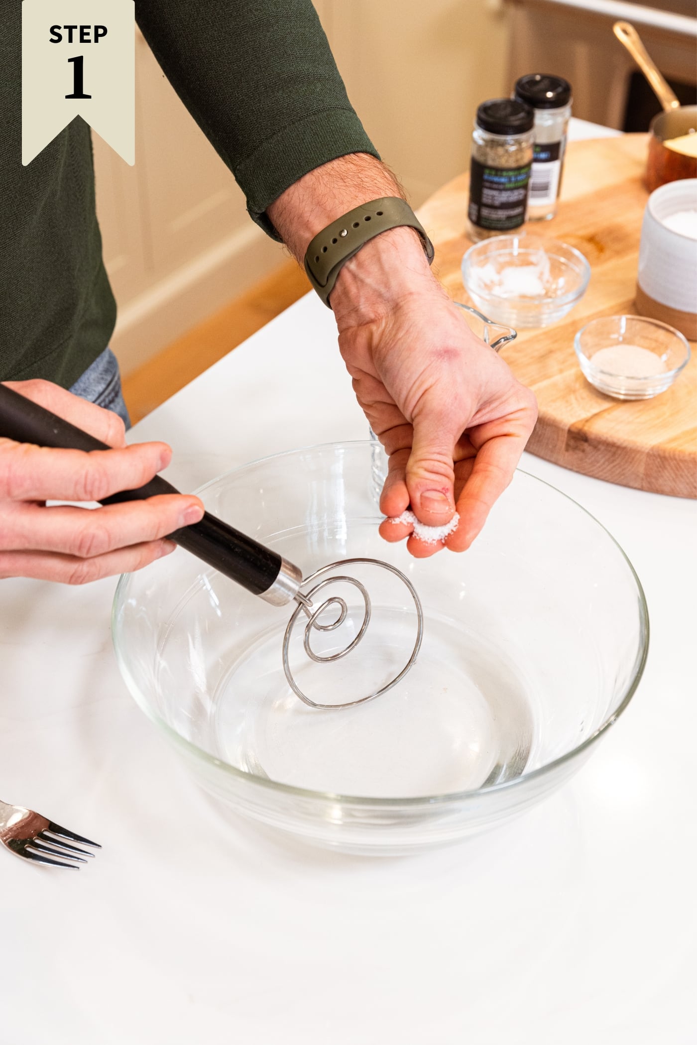Large glass bowl on white countertop with water in bottom with hand stirring together mixture with wire whisk.