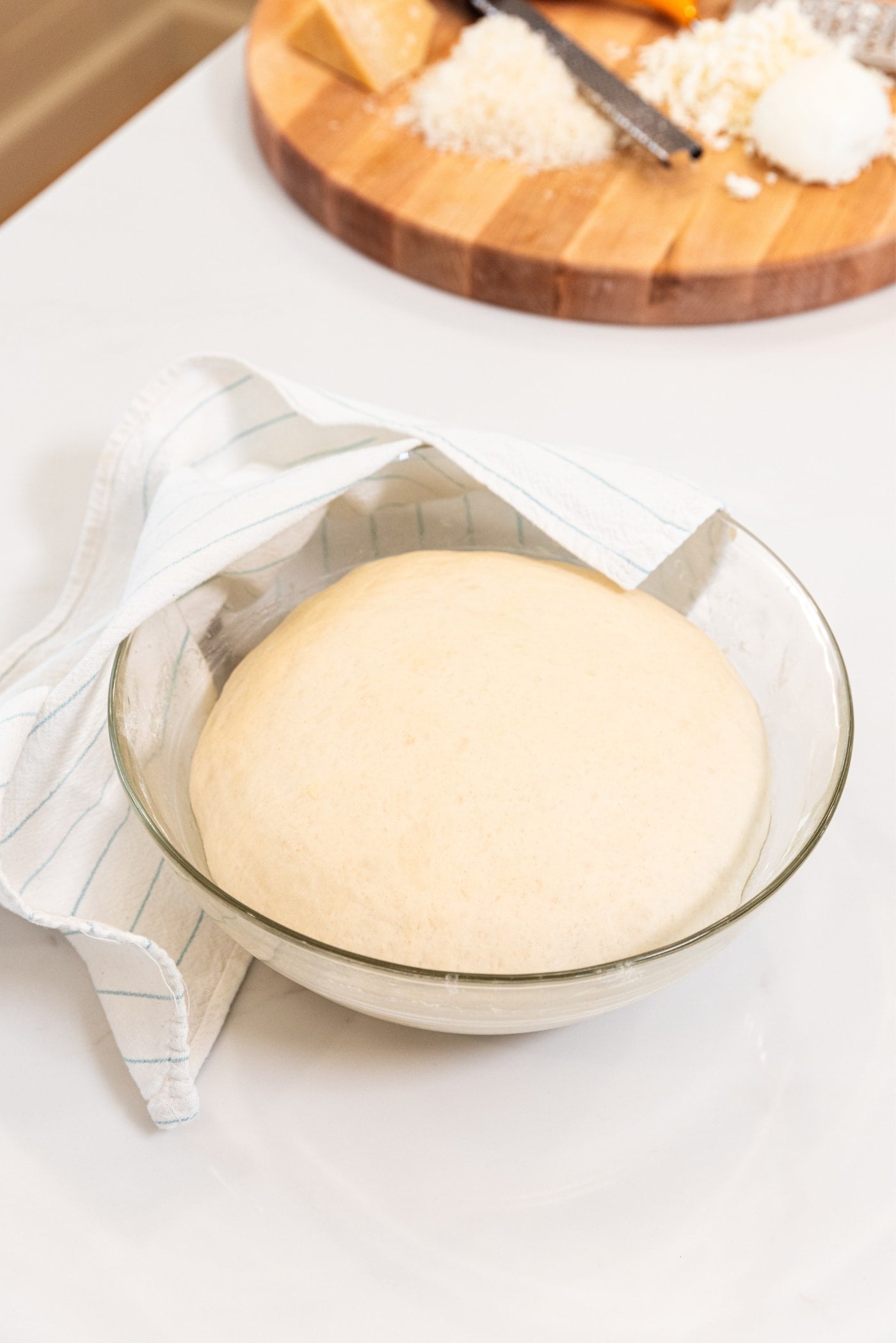 White surface with large glass bowl with dough risen inside with towel resting on side of the bowl.