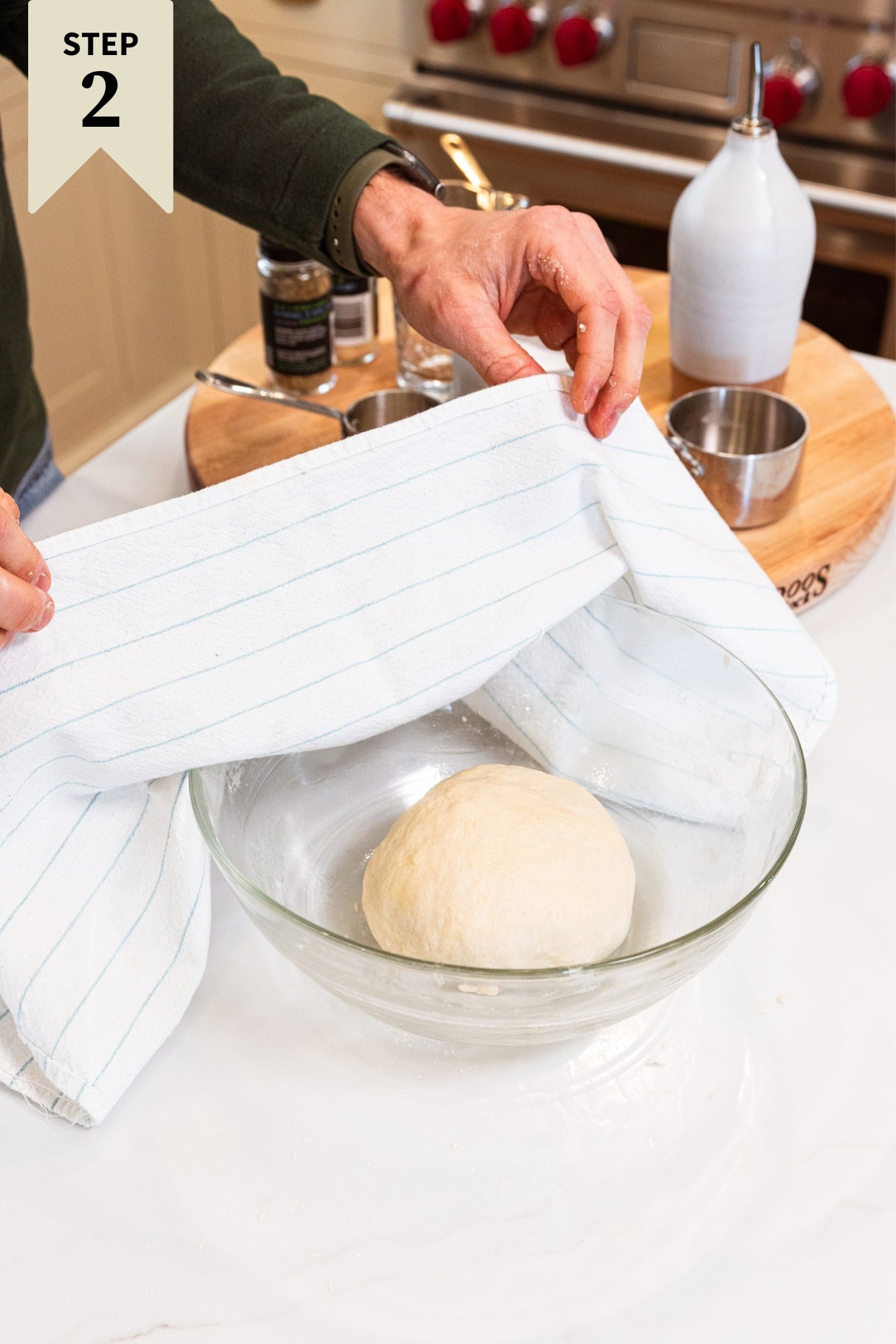 Hand holding cloth towel putting over glass bowl on white countertop with dough inside bowl before the dough rises in the bowl.