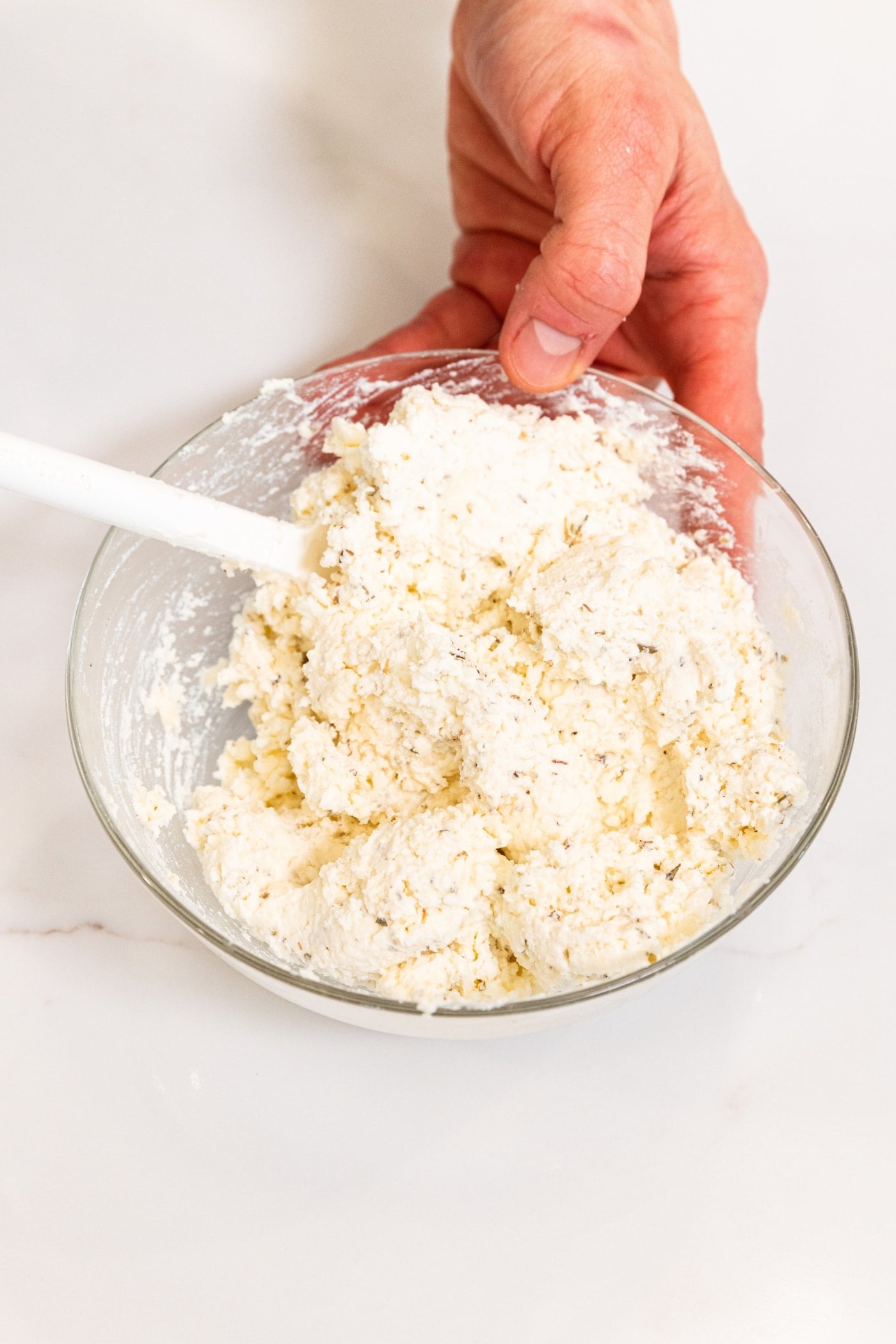 Hand using white spatula to mix together three cheeses in a small glass bowl on white surface.