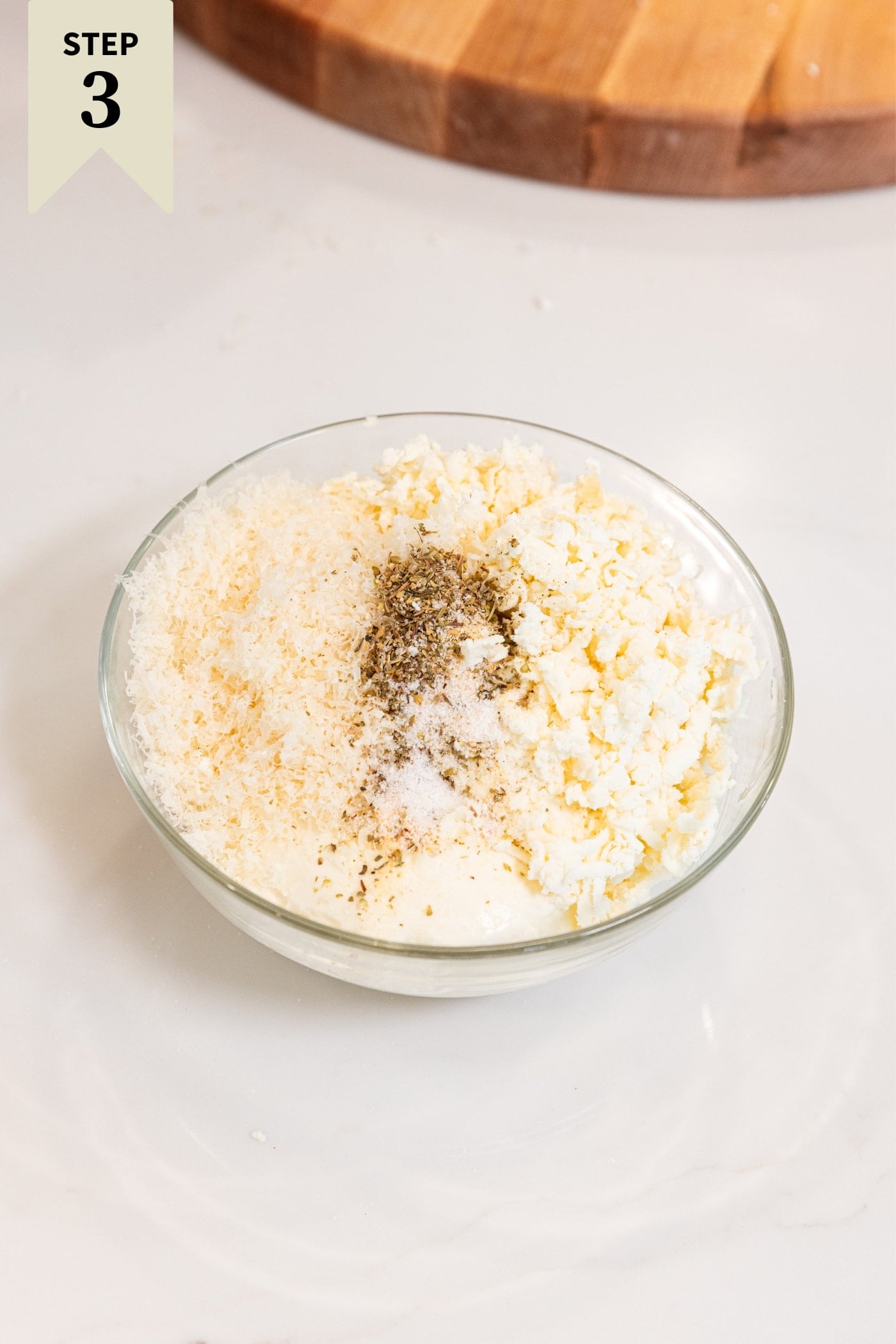 Medium-sized glass bowl sitting on white countertop filled with ricotta cheese, shredded mozzarella, and Parmesan cheese with spices on top right before being mixed together.