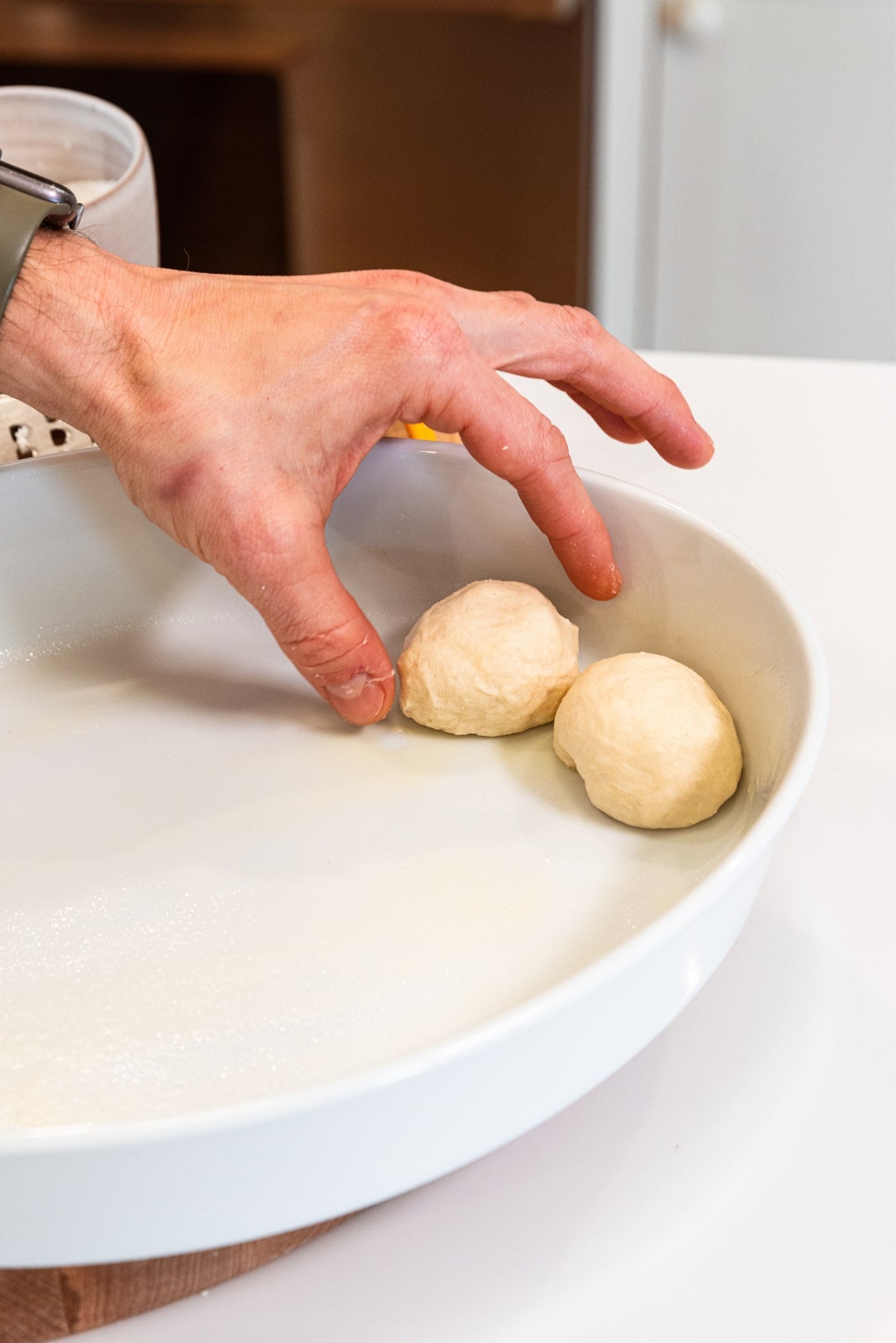 Hand placing dough ball in oval metal baking dish with one other dough ball as well.