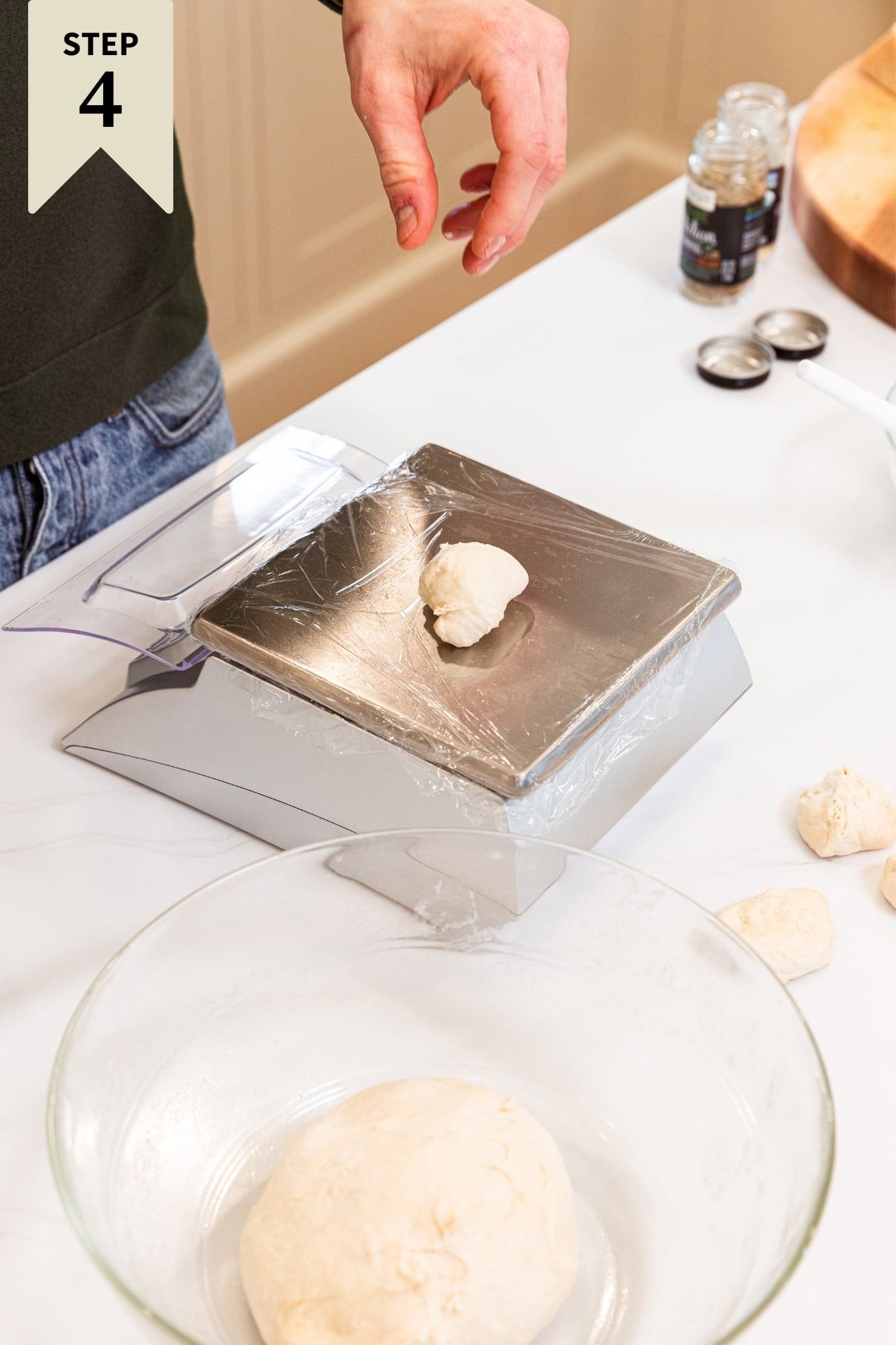 Food scale with small piece of dough sitting on surface with rest of dough in glass bowl to the side all on white countertop.