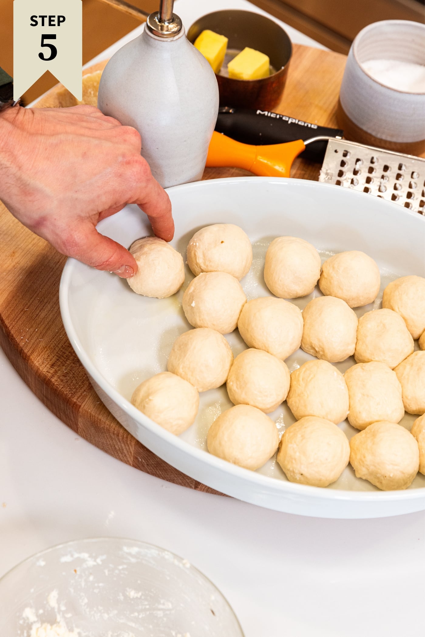 Hand placing pizza bite into an oval baking dish already filled with lots of other pizza bites on wood cutting board before rising for a second time.